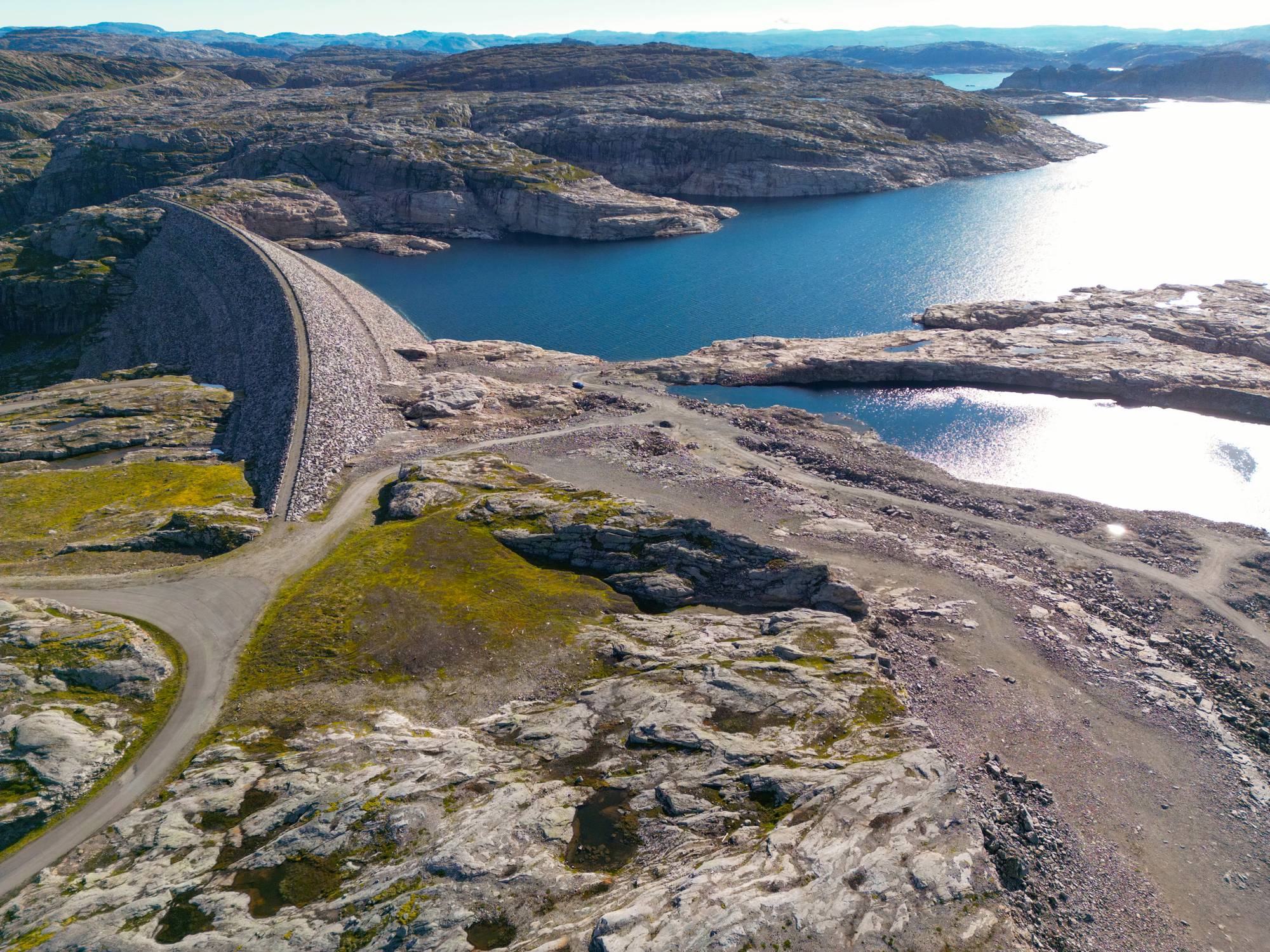 Et stort steinfyllingsdam. Vann fyller reservoaret bak demningen. Omgivelsene består av steinete landskap med noe vegetasjon. (Bildebeskrivelsen er laget av en KI-tjeneste)