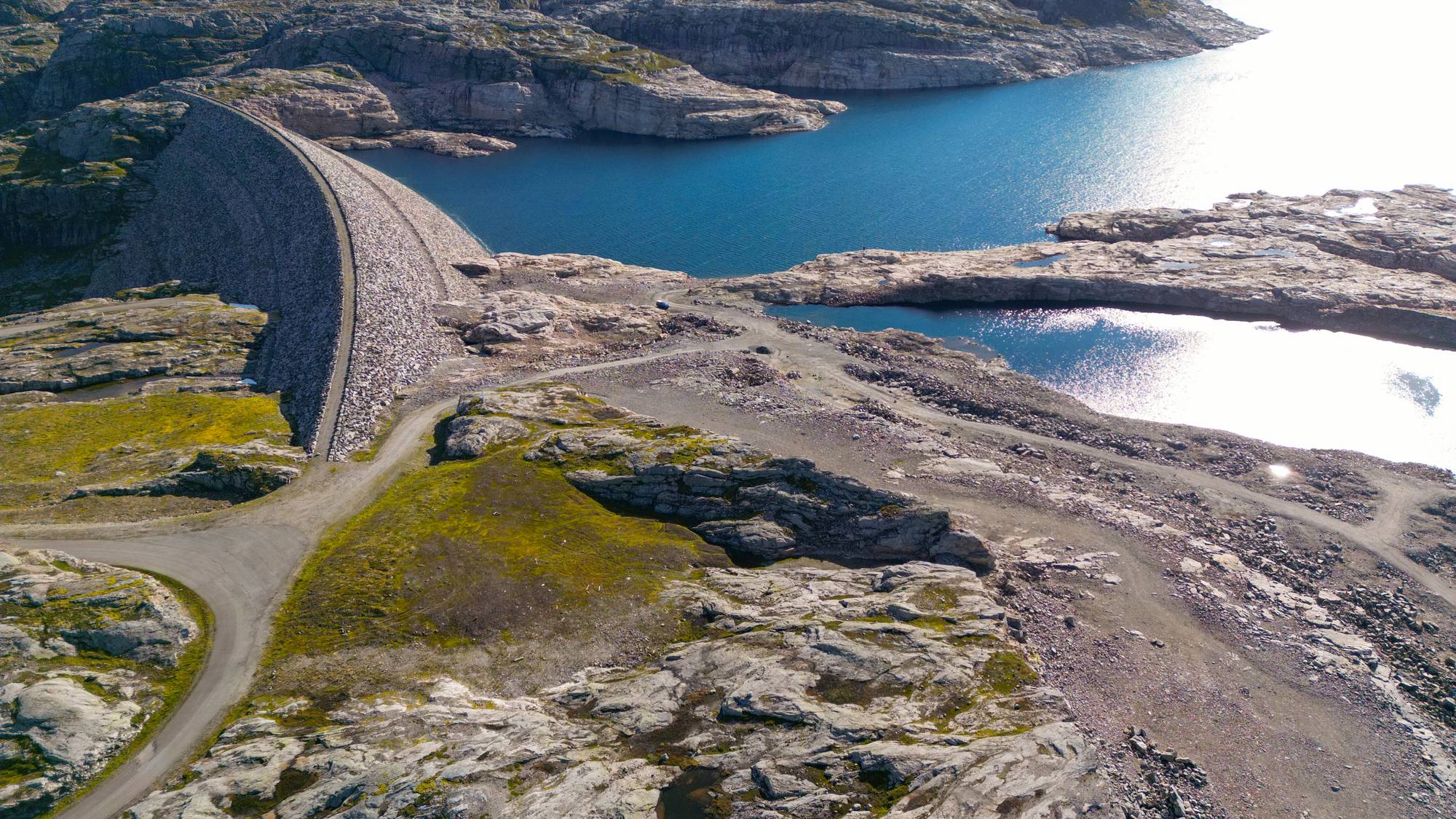 Et stort steinfyllingsdam. Vann fyller reservoaret bak demningen. Omgivelsene består av steinete landskap med noe vegetasjon. (Bildebeskrivelsen er laget av en KI-tjeneste)