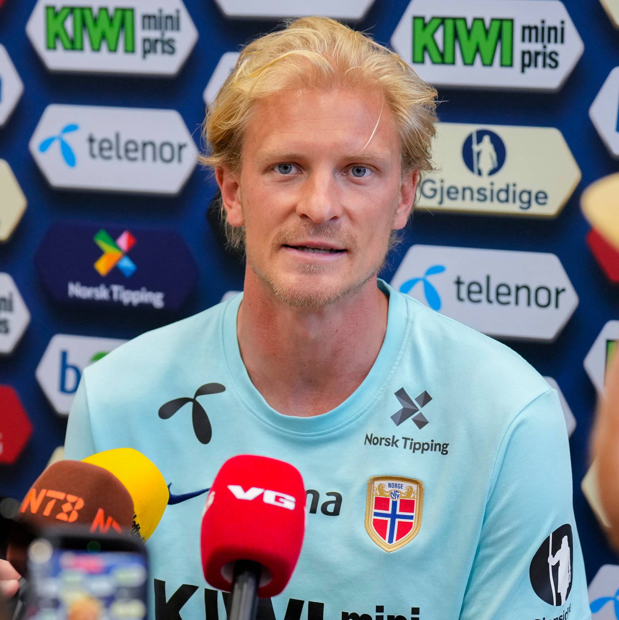 En mann med blondt hår sitter og snakker til tre mikrofoner. Bak ham er en vegg med logoer fra flere sponsorer. Foran mannen er det tre mobiltelefoner som filmer ham. (Bildebeskrivelsen er laget av en KI-tjeneste)