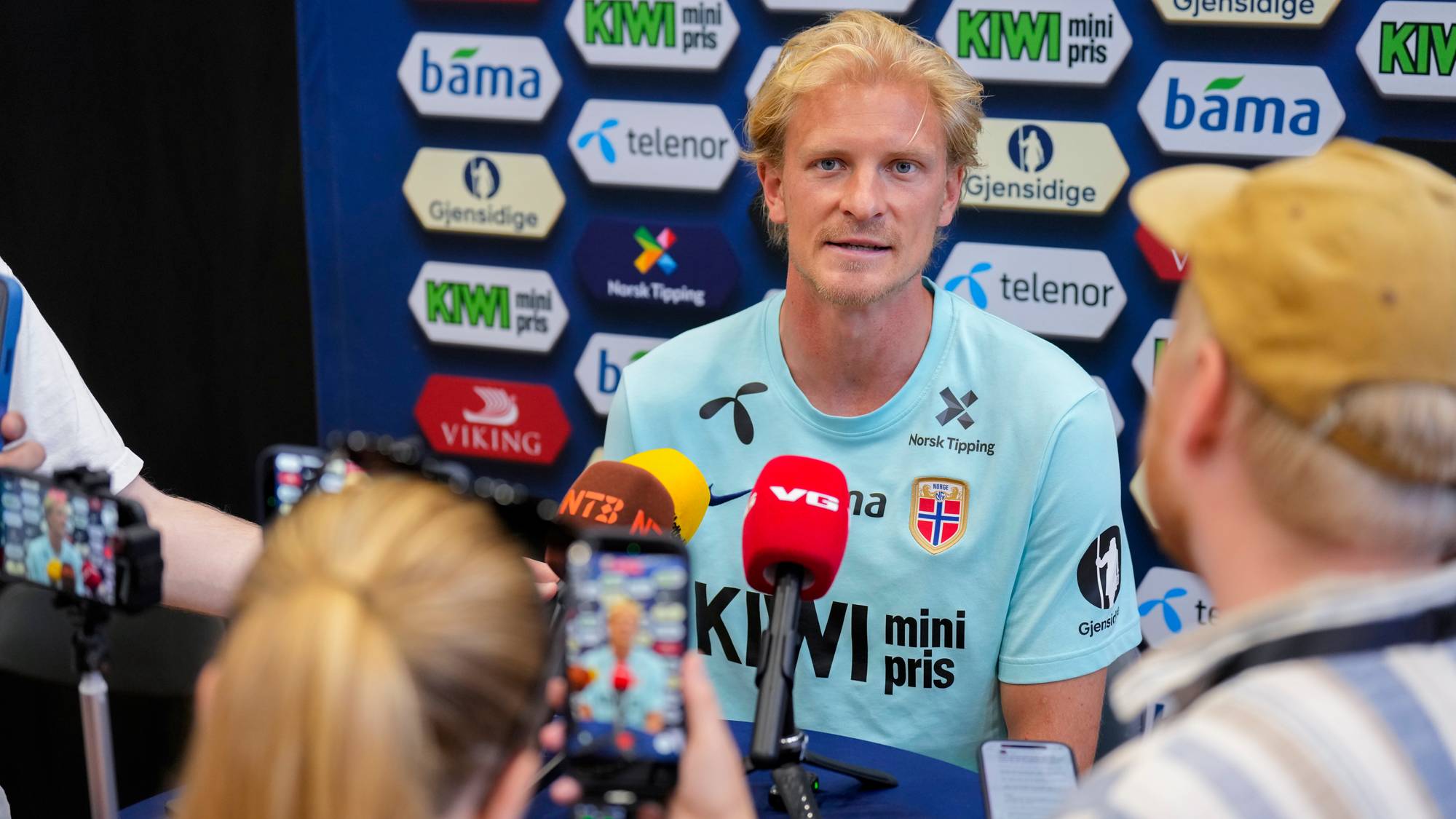 En mann med blondt hår sitter og snakker til tre mikrofoner. Bak ham er en vegg med logoer fra flere sponsorer. Foran mannen er det tre mobiltelefoner som filmer ham. (Bildebeskrivelsen er laget av en KI-tjeneste)