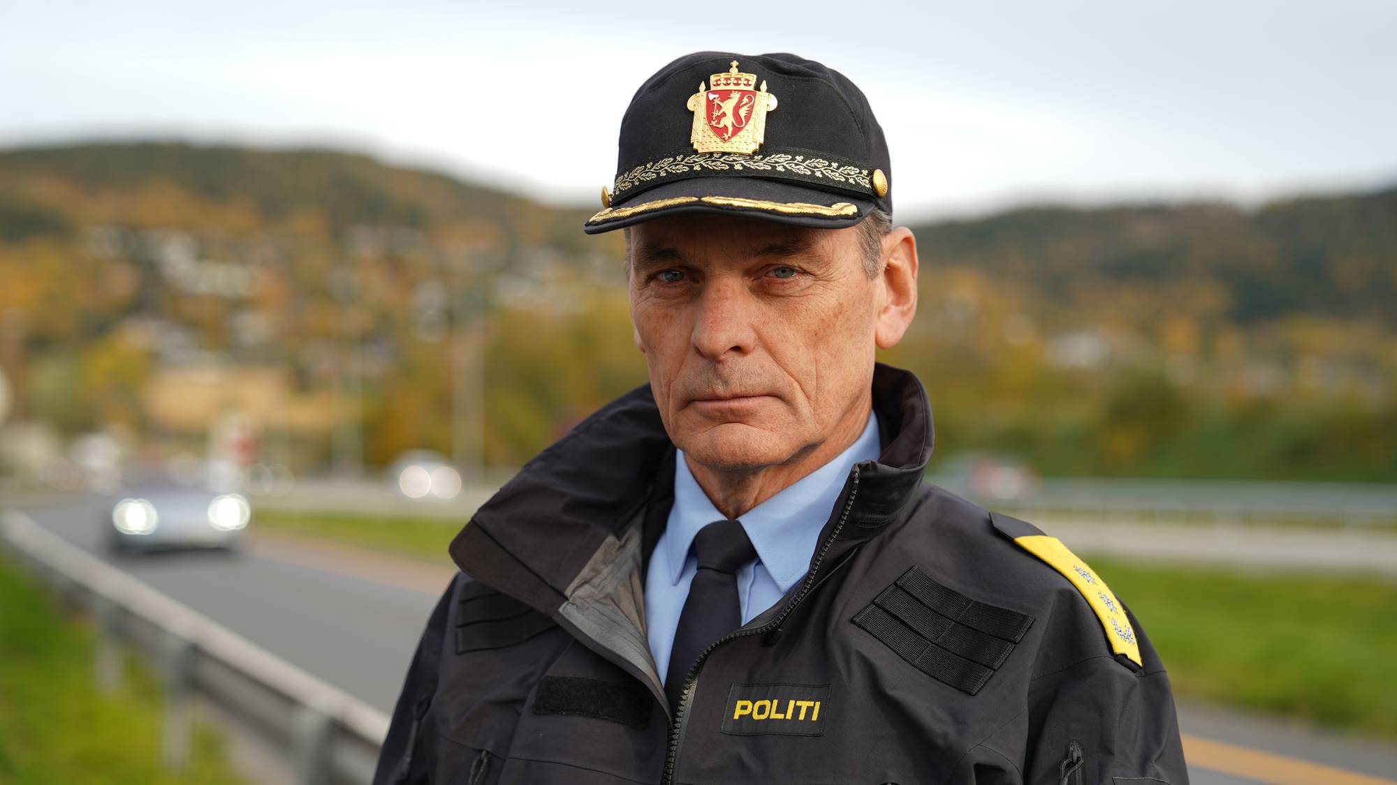 Portrett av en politimann. Han bærer uniform og caps. I bakgrunnen er det en vei med en bil, og et grønt, høstfarget landskap med trær. (Bildebeskrivelsen er laget av en KI-tjeneste)