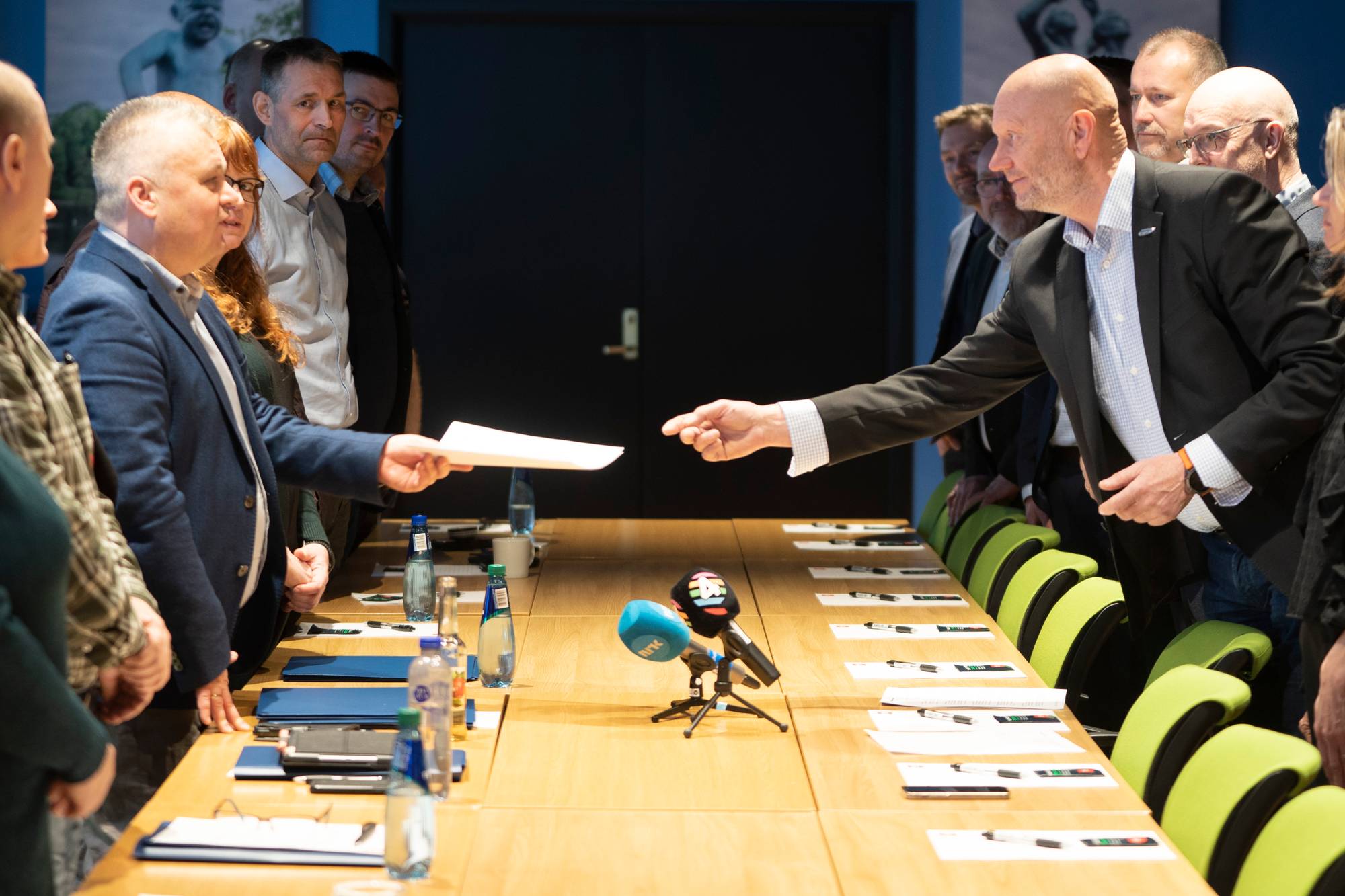 En mann i mørk dress rekker et dokument over et langt bord til en annen mann. Begge er i profil og de andre møtedeltakerne er synlige bak dem. Møterommet har en mørk bakgrunn og grønne stoler langs bordet. (Bildebeskrivelsen er laget av en KI-tjeneste)