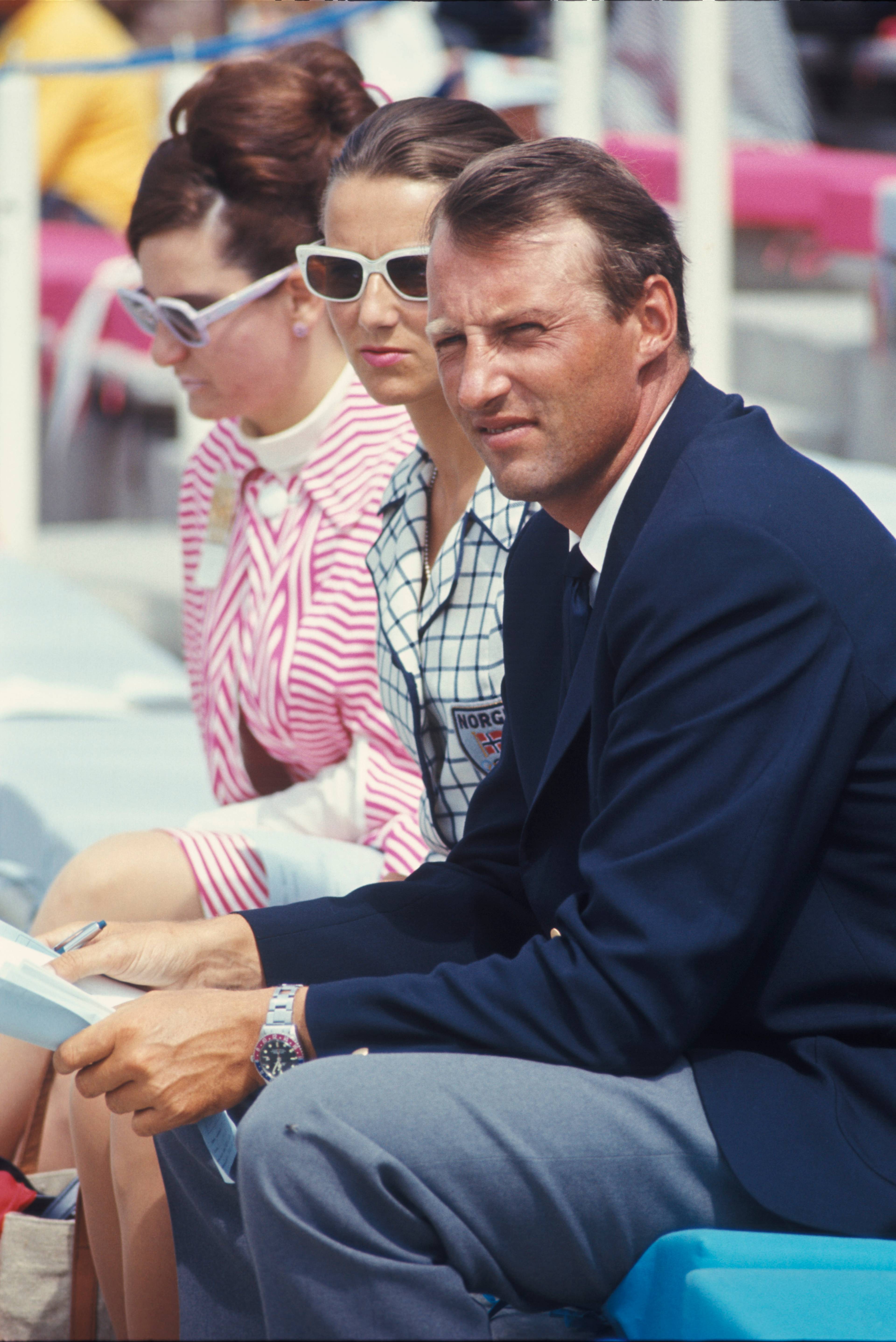 Kong Harald og Dronning Sonja sitter på en tribune, ved siden av en kvinne, under et arrangement. (Bildebeskrivelsen er laget av en KI-tjeneste)