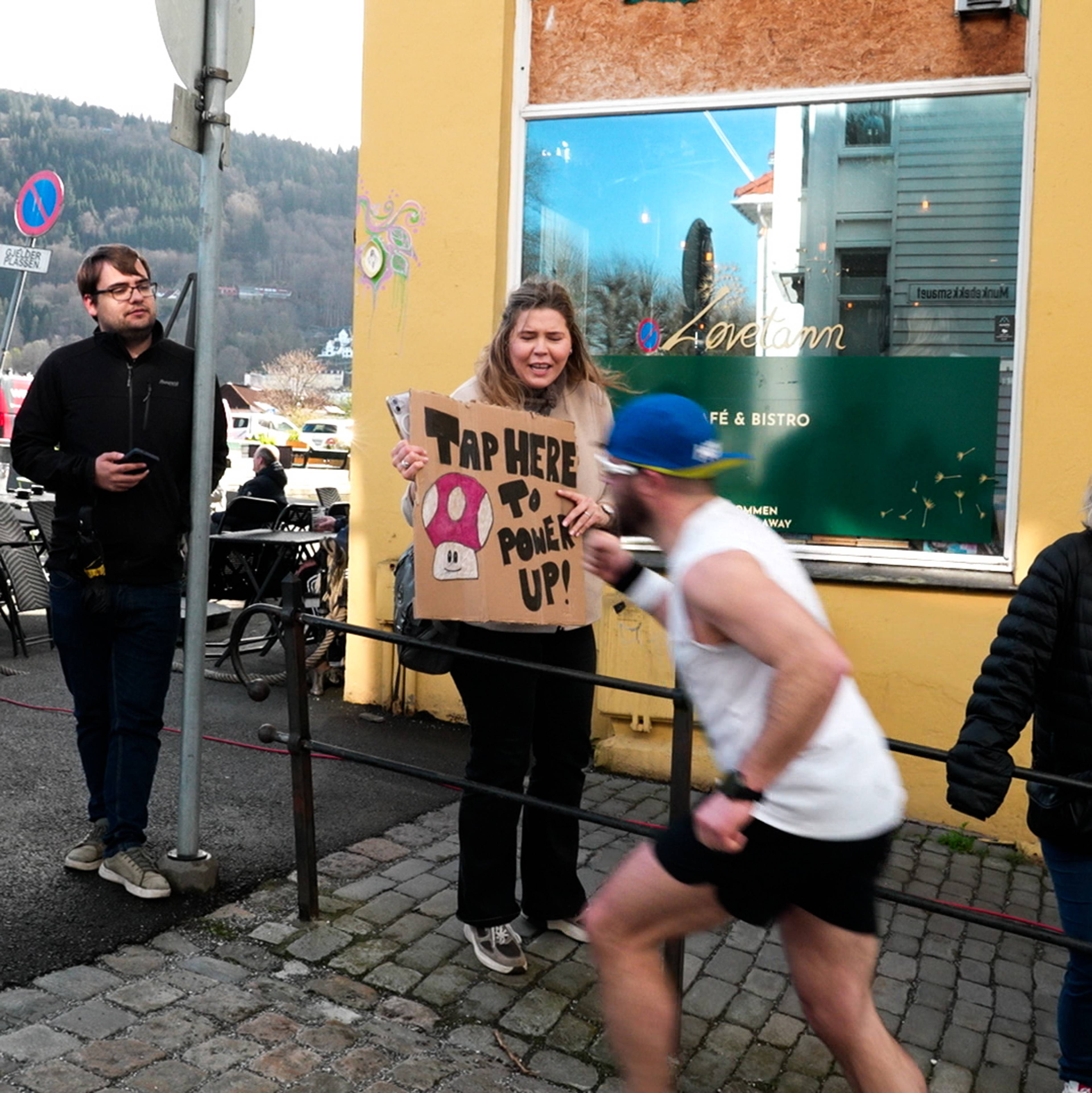 Tilskodar med oppmuntrande plakat langs løypa på Bergen City Marathon 2026: "Tap here to power up".