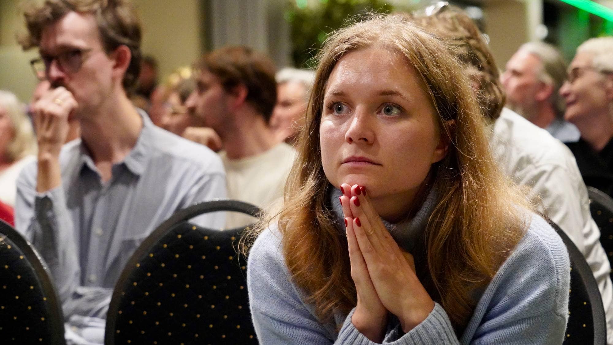 Kvinne med hendene samlet foran ansiktet sitter i en forsamling. Flere personer sitter bak henne. (Bildebeskrivelsen er laget av en KI-tjeneste)