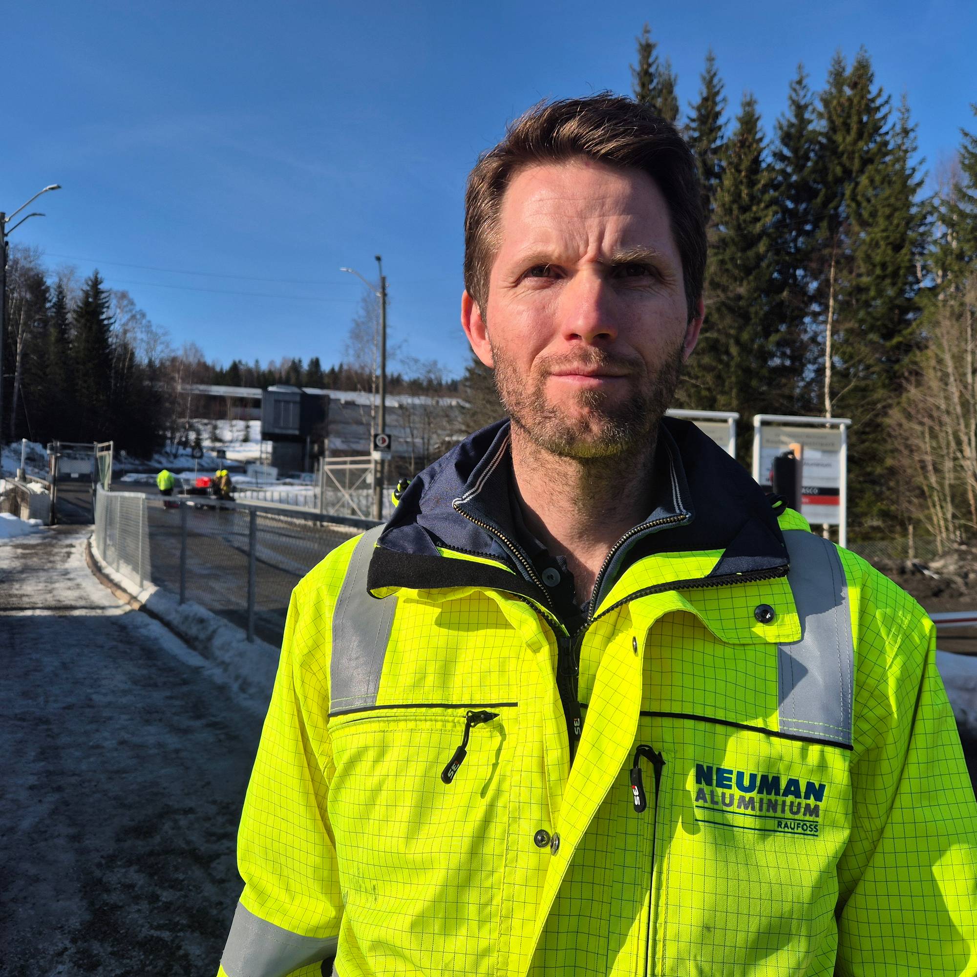 En person iført en neon gul arbeidsjakke står foran et inngangsområde. Bak personen er det snødekte områder og et gjerde som fører til en bro. I bakgrunnen er det flere personer i arbeid samt en innsjø. Lysforholdene er gode, og det er klart vær. (Bildebeskrivelsen er laget av en KI-tjeneste)