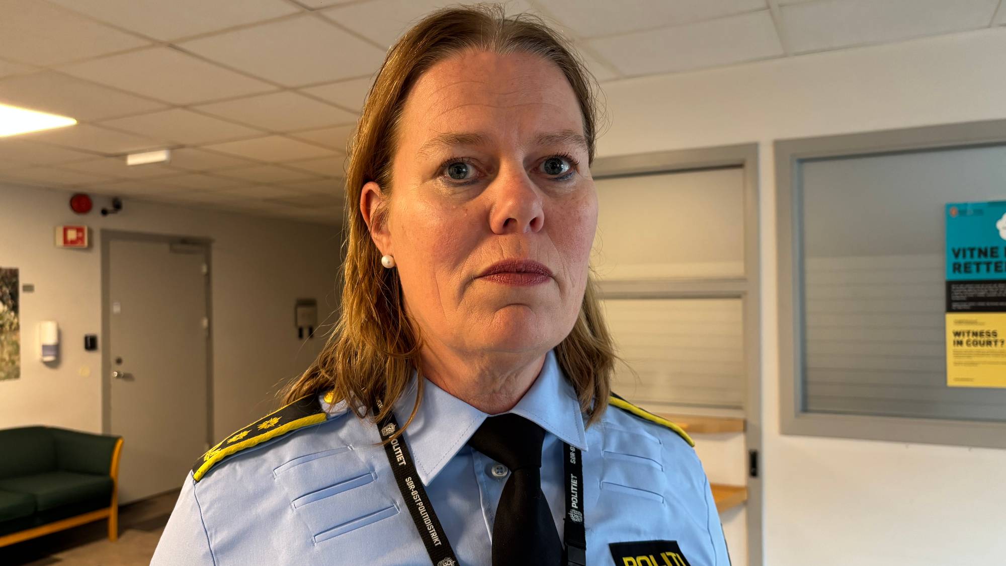 En kvinnelig politibetjent står i en offentlig bygning, iført uniform og ID-kort. Omgivelsene inkluderer en sofa og informasjonstavler på veggene. Det er lav belysning og et industrielt preg. Betjenten ser mot kameraet og har langt hår.