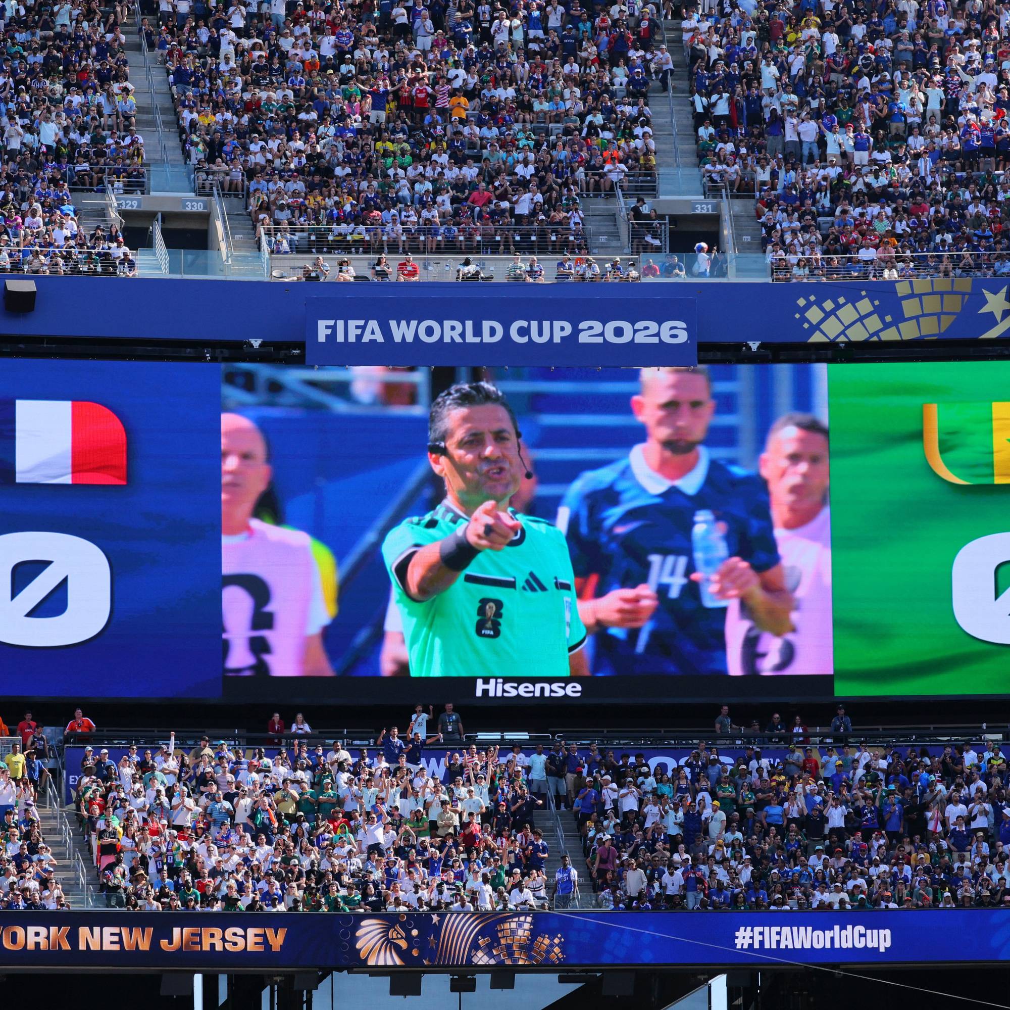 Storskjerm viser resultattavle for fotballkampen mellom Frankrike og Senegal. En dommer med grønn trøye peker mot tilskuerne. Tribune med mye publikum i bakgrunnen. FIFA World Cup 2026-logo vises på skjermen. (Bildebeskrivelsen er laget av en KI-tjeneste)