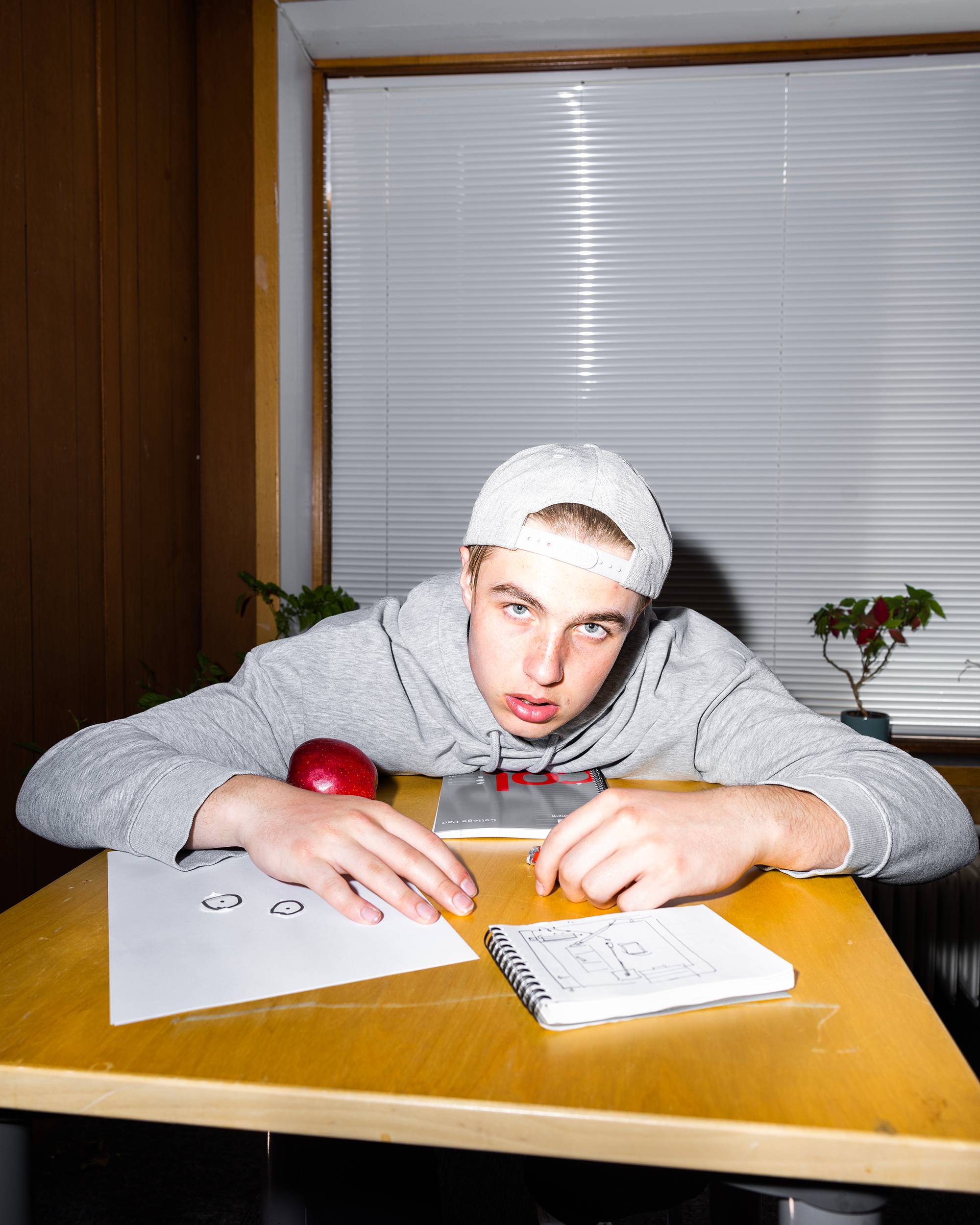 Vetle er kledd ut som skoleelev. Han har på seg caps og hettegenser. Han lener seg over pulten med et frustrert blikk. På pulten er det et eple og skrivesaker. 
