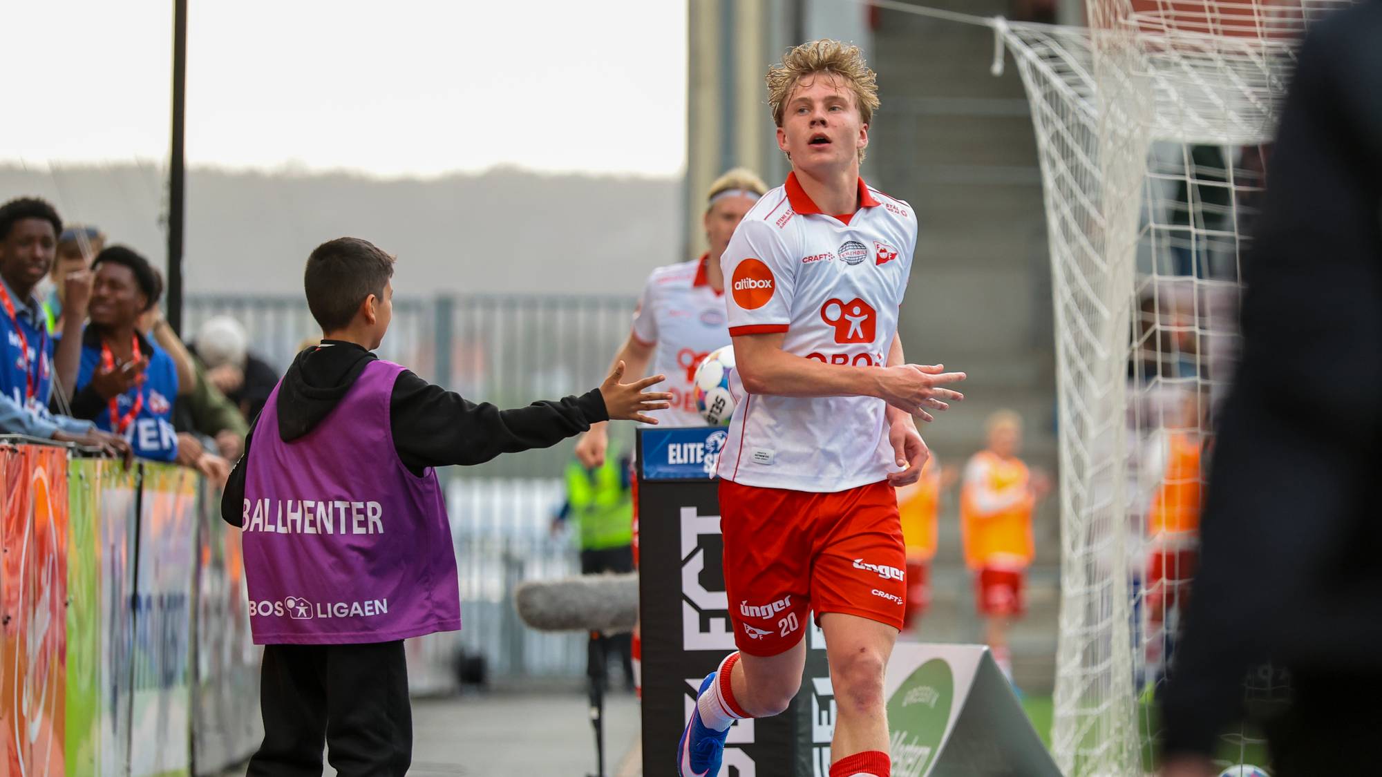 En fotballspiller i hvit og rød drakt løper på en bane. En gutt i lilla vest med teksten BALLHENTER står ved siden av. Tilskuere ser på fra venstre. (Bildebeskrivelsen er laget av en KI-tjeneste)