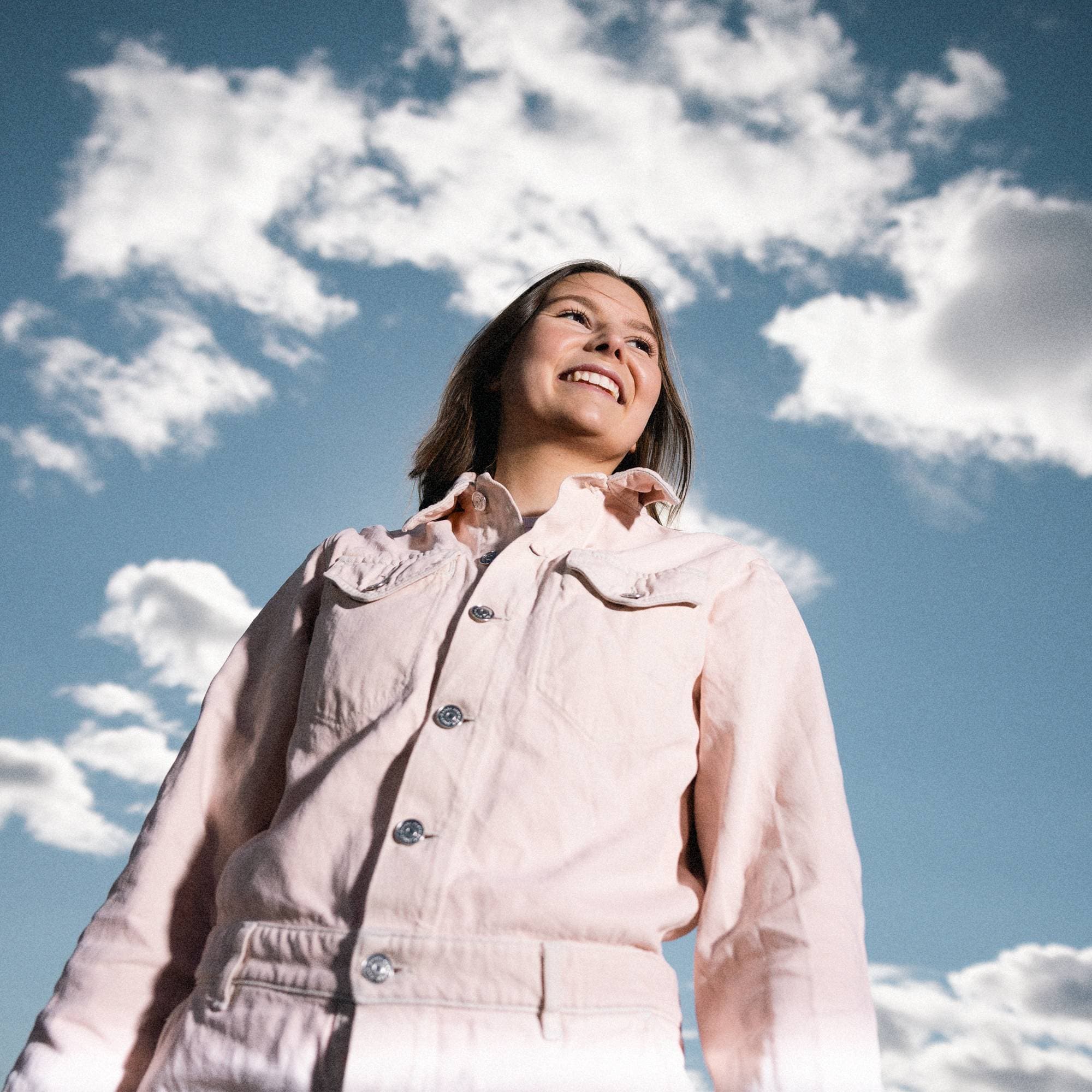 Bilde av Hedda, som står i rosa klær foran blå himmel med skyer. 