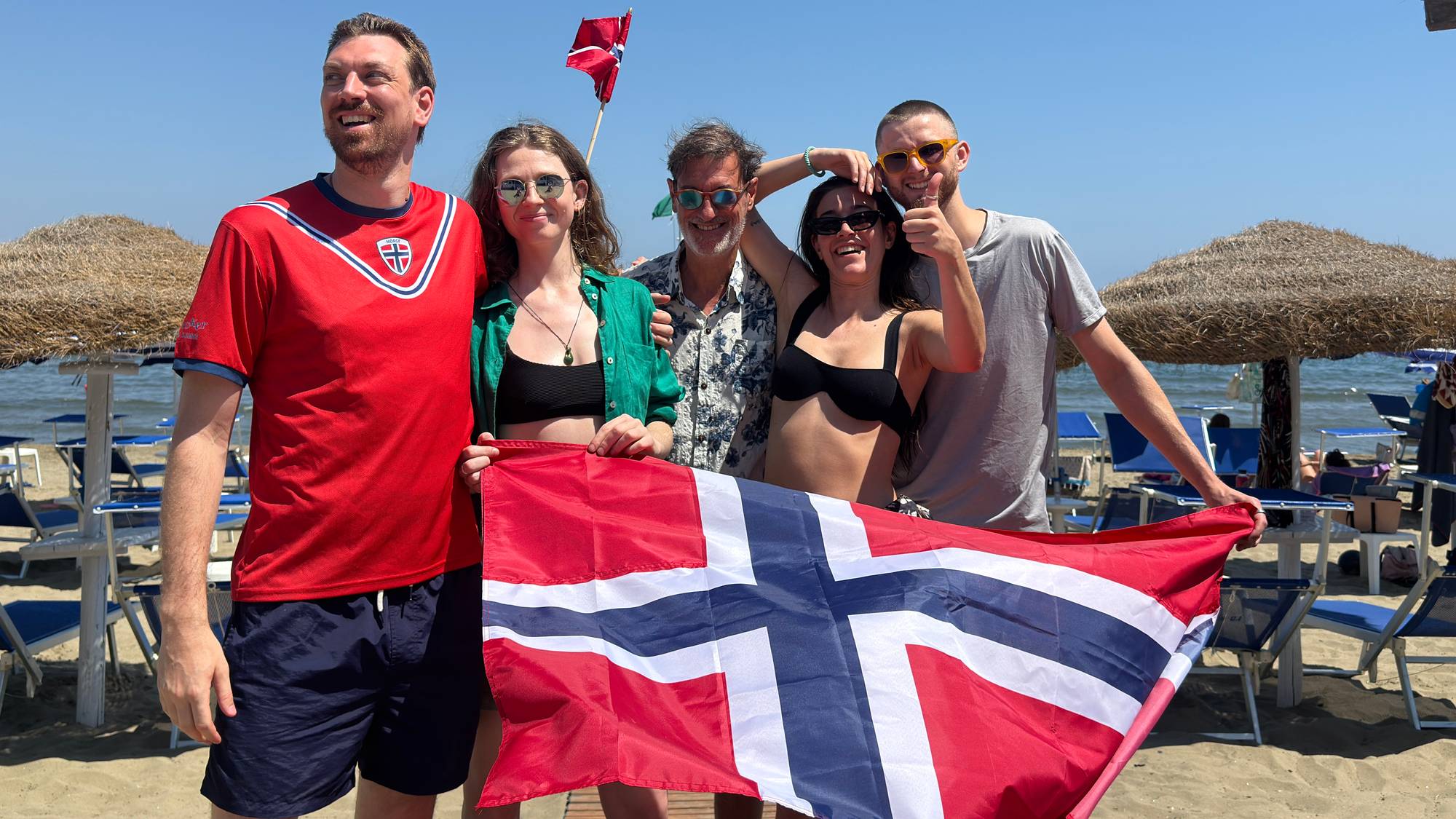 Bildet viser en gruppe mennesker på en strand med et stort norsk flagg.
