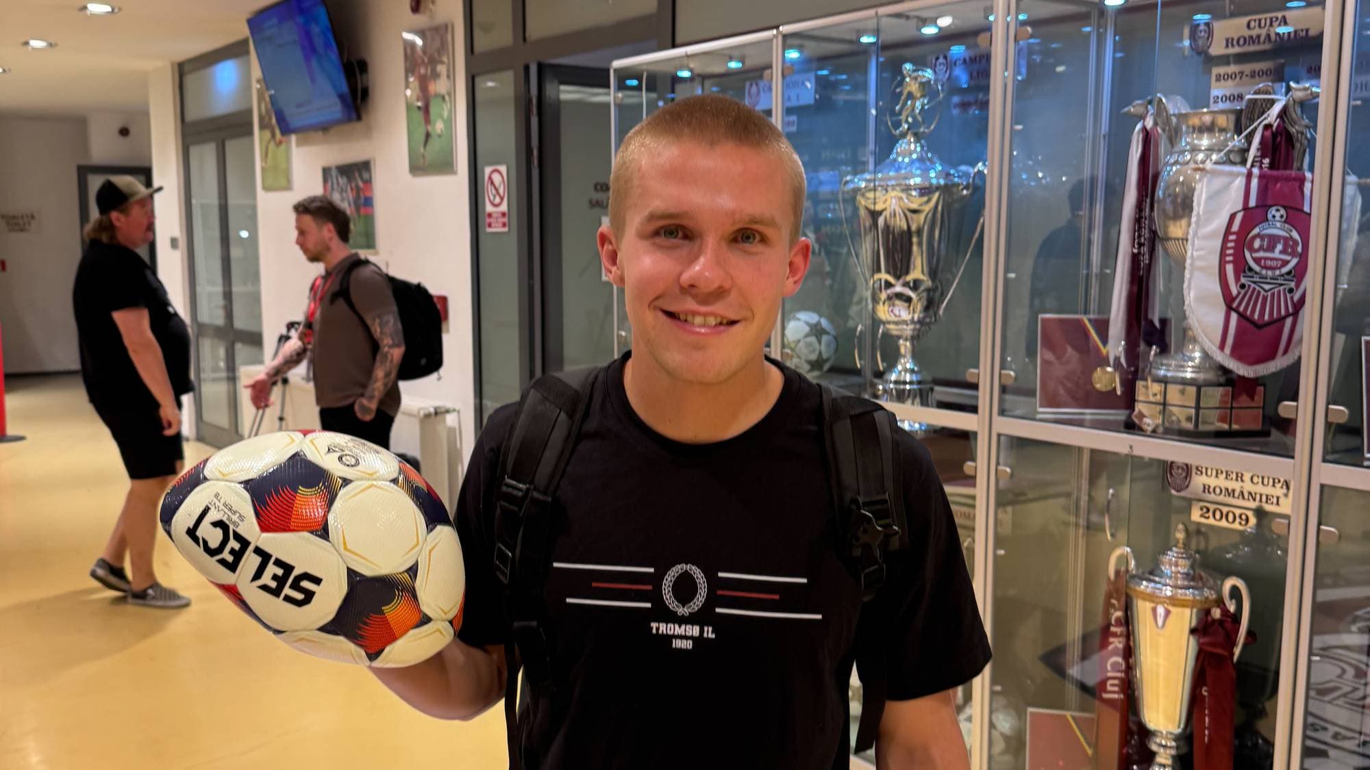 Ung mann holder en ball foran en montre med pokaler i en korridor. (Bildebeskrivelsen er laget av en KI-tjeneste)