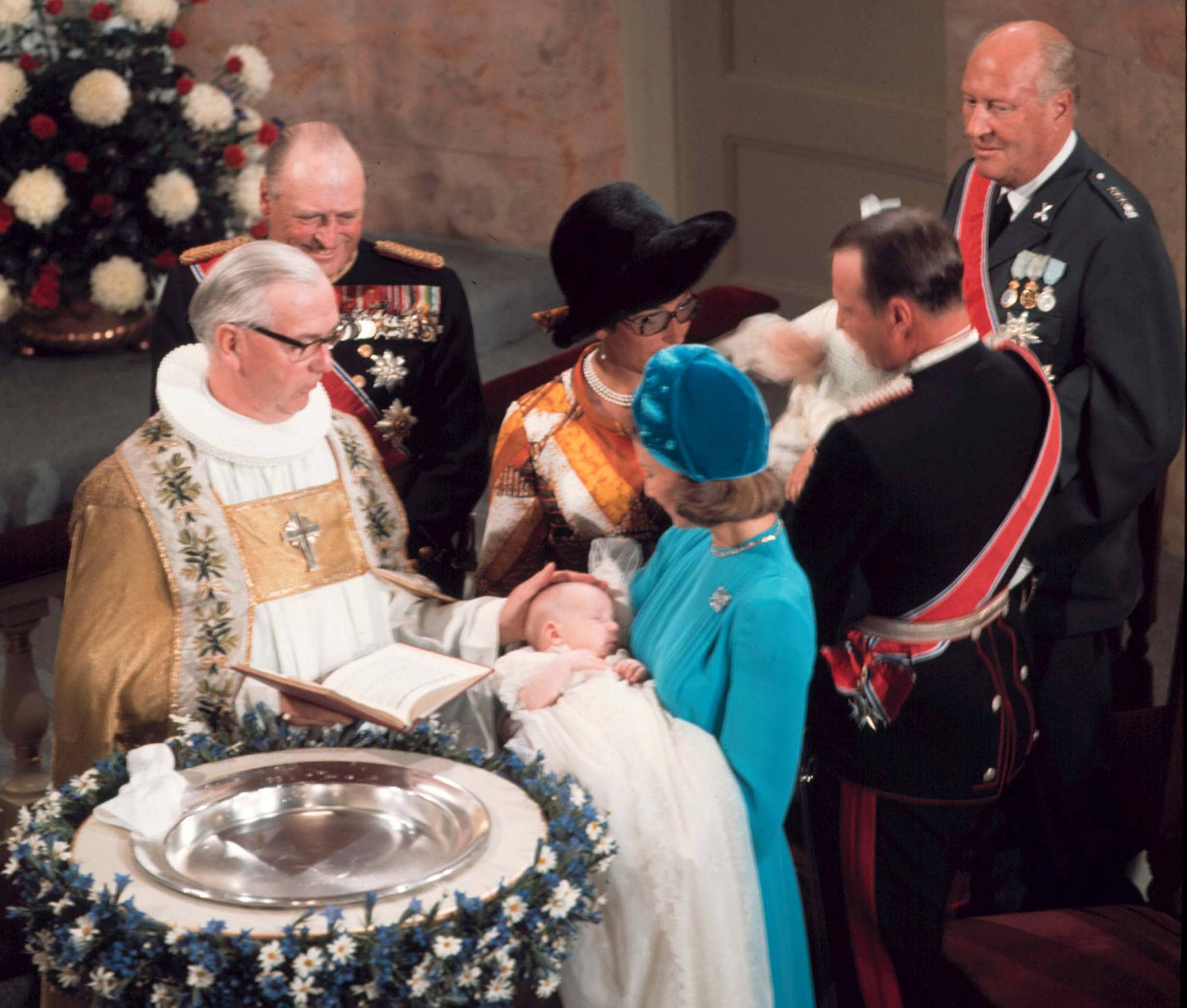 Biskopen legger hånden på dåpsbarnet prins Haakon Magnus' hode, mens kronprinsesse Sonja holder barnet ved døpefonten. Bak henne står kronprins Harald med prinsesse Märtha Louise på armen, og ved siden av står prinsesse Astrid og kong Olav.