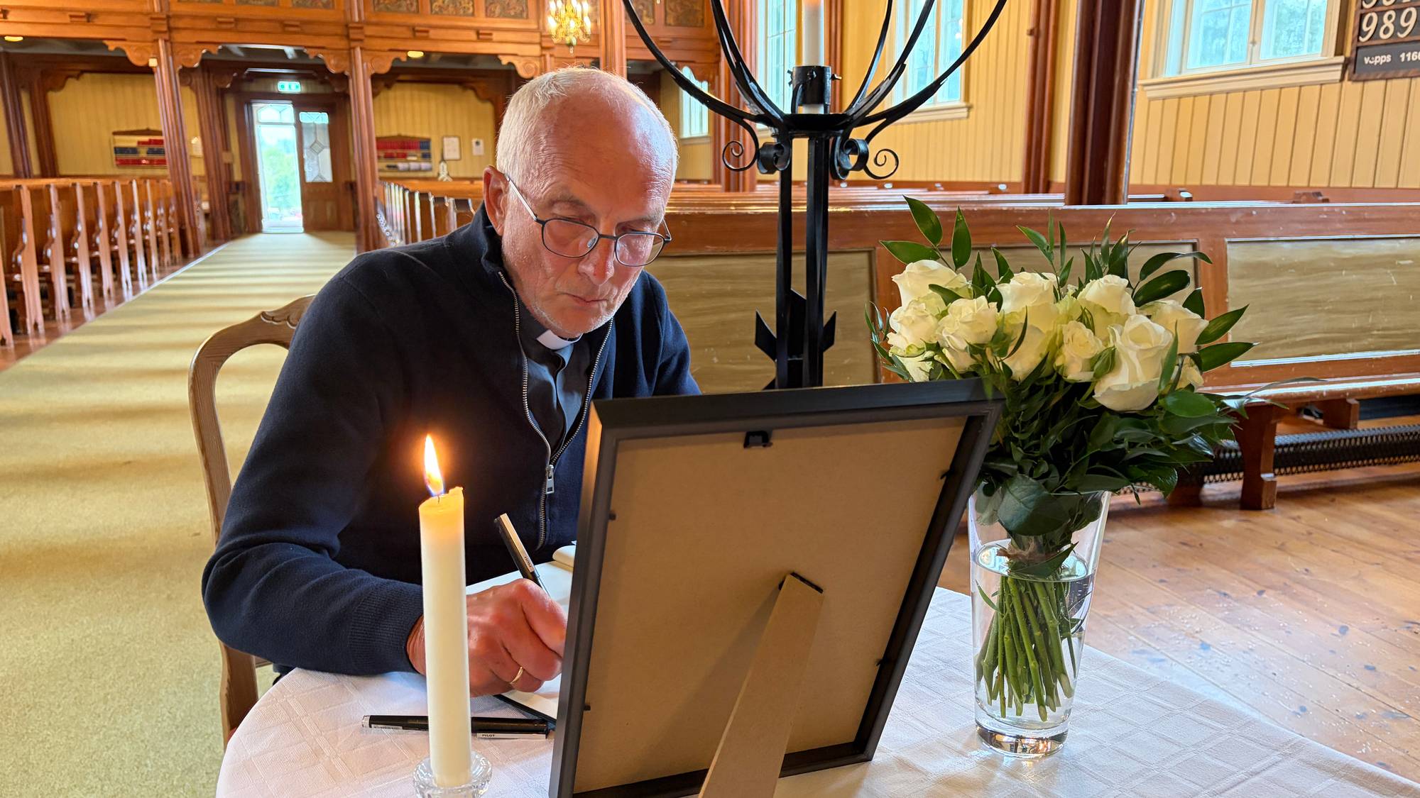 En prest skriver i en minneprotokoll ved et bord i en kirke. Det står et bilde og hvite roser der. 