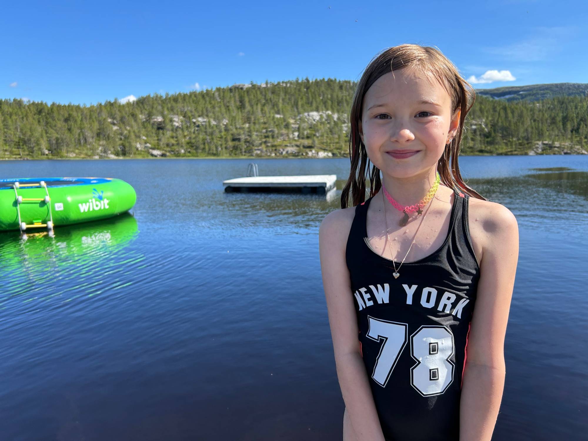Ebba Strøm Mathisen står på bryggekanten ved en innsjø i en dryppende våt, sort badedrakt der det står New York og tallet 78. Bak henne flyter en vanntrampoline og en flytebrygge. 
