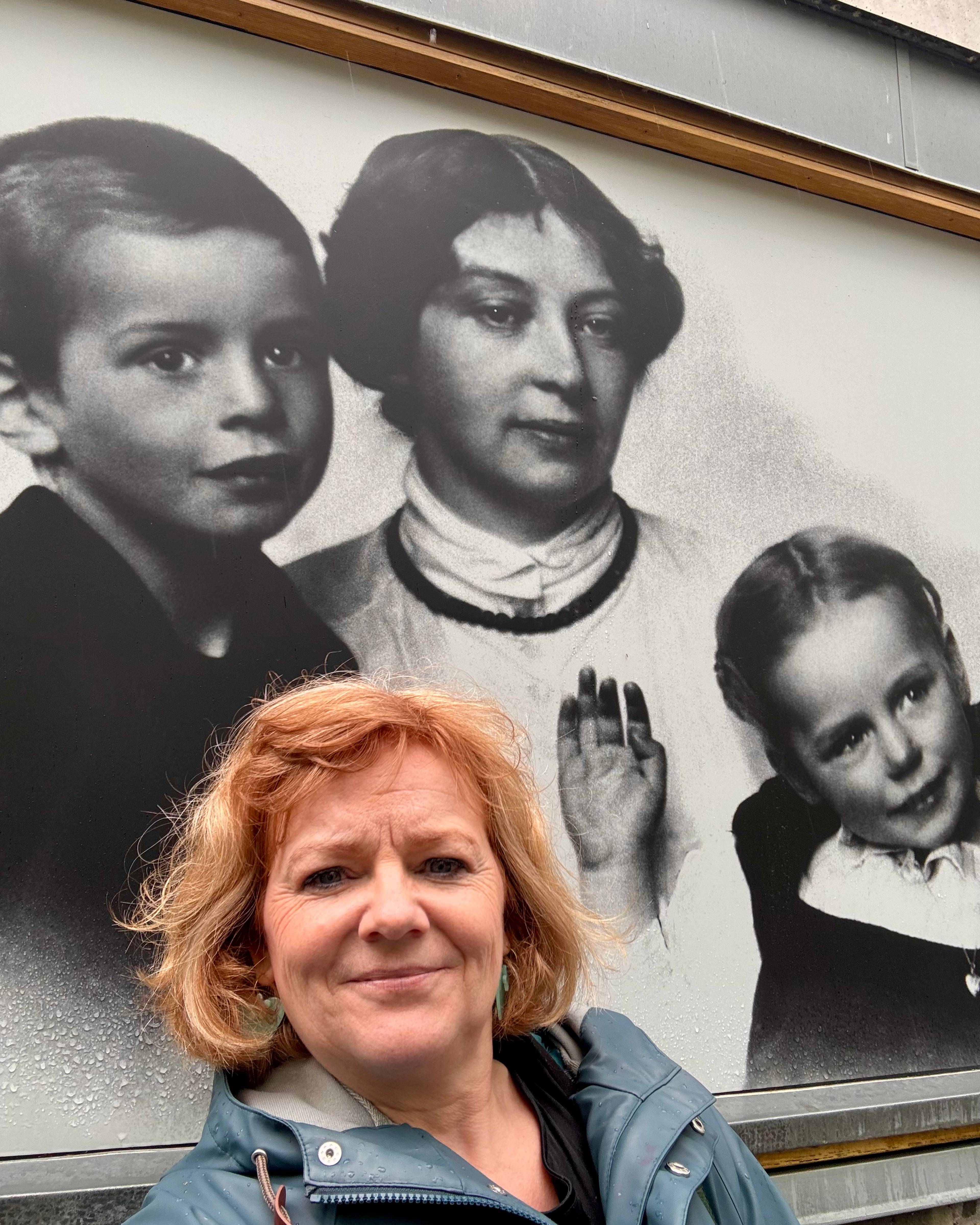 En stort bilde i svart-hvitt på en vegg med Sigrid Undset og barna. Foran står en blond kvinne i regnfrakk