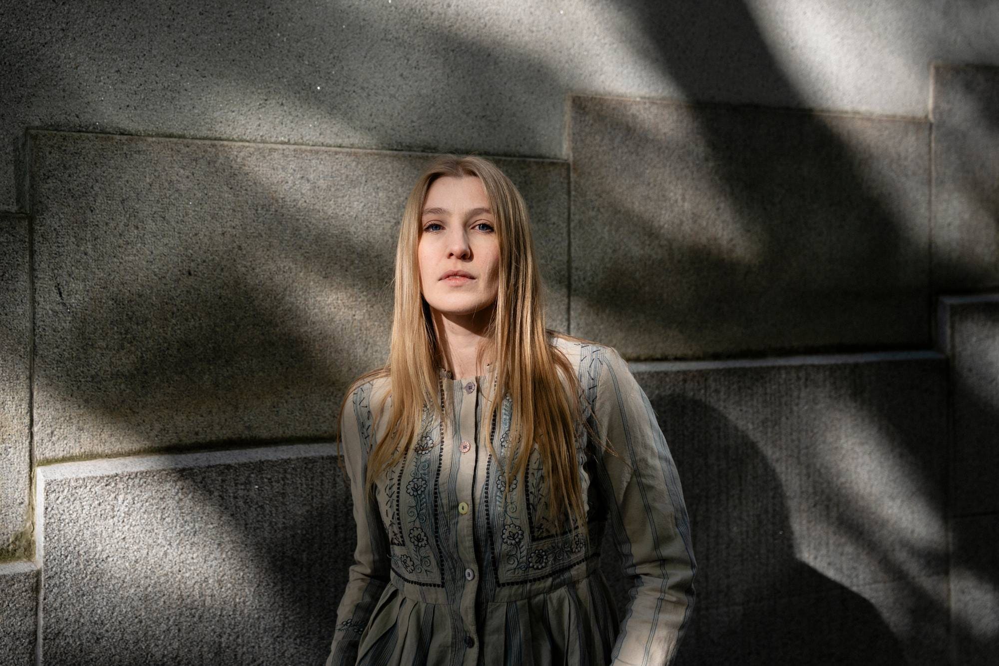 En kvinne med langt, blondt hår står foran en steinvegg. Hun har på seg en beige kjole med blått mønster og striper. Kvinnen ser mot kameraet. Skygger faller fra en struktur over og til venstre for henne. (Bildebeskrivelsen er laget av en KI-tjeneste)