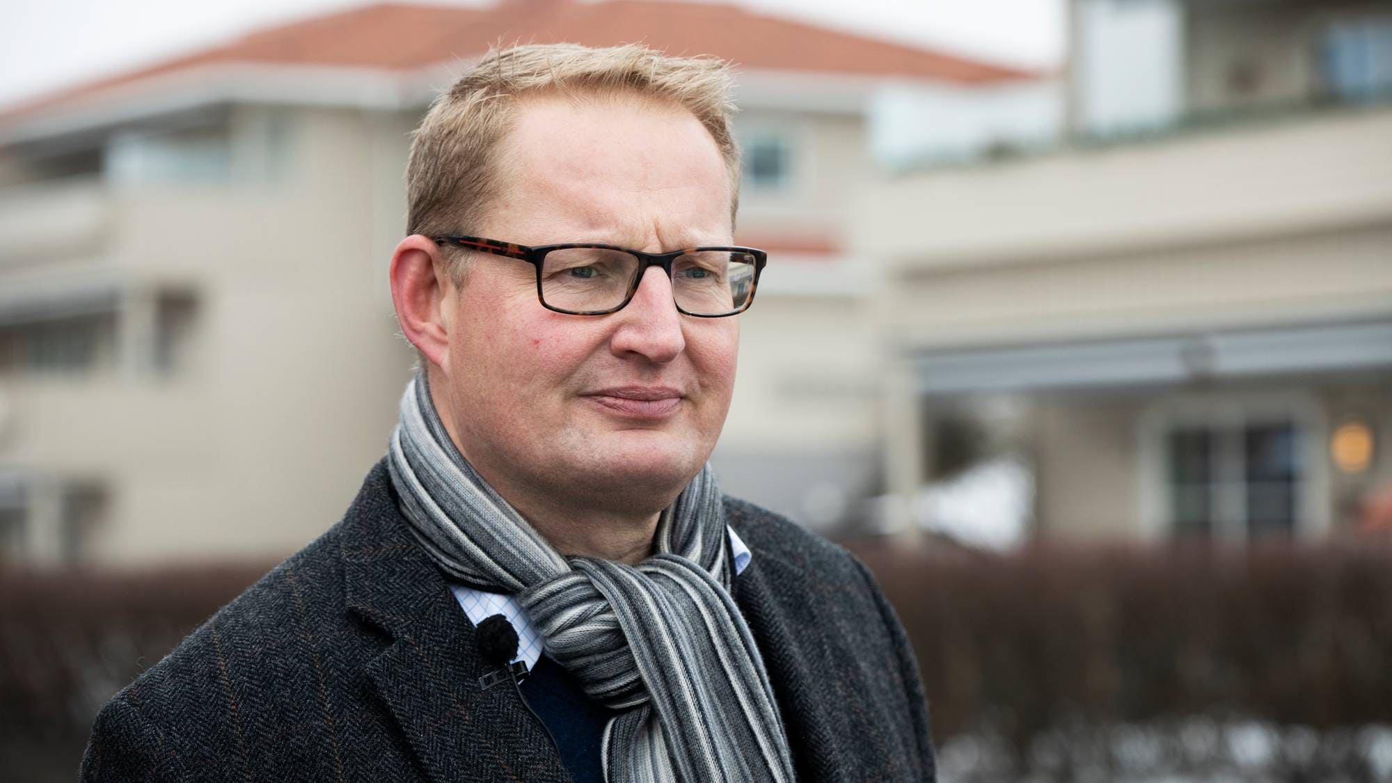En mann med briller og skjerf står foran en bygning. Han har på seg en mørk ytterjakke og ser mot kameraet. I bakgrunnen er det flere bygninger og busker som delvis skjuler utsikten. Bildet er tatt utendørs på en grå dag.