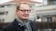 Anders Fehn En mann med briller og skjerf står foran en bygning. Han har på seg en mørk ytterjakke og ser mot kameraet. I bakgrunnen er det flere bygninger og busker som delvis skjuler utsikten. Bildet er tatt utendørs på en grå dag.