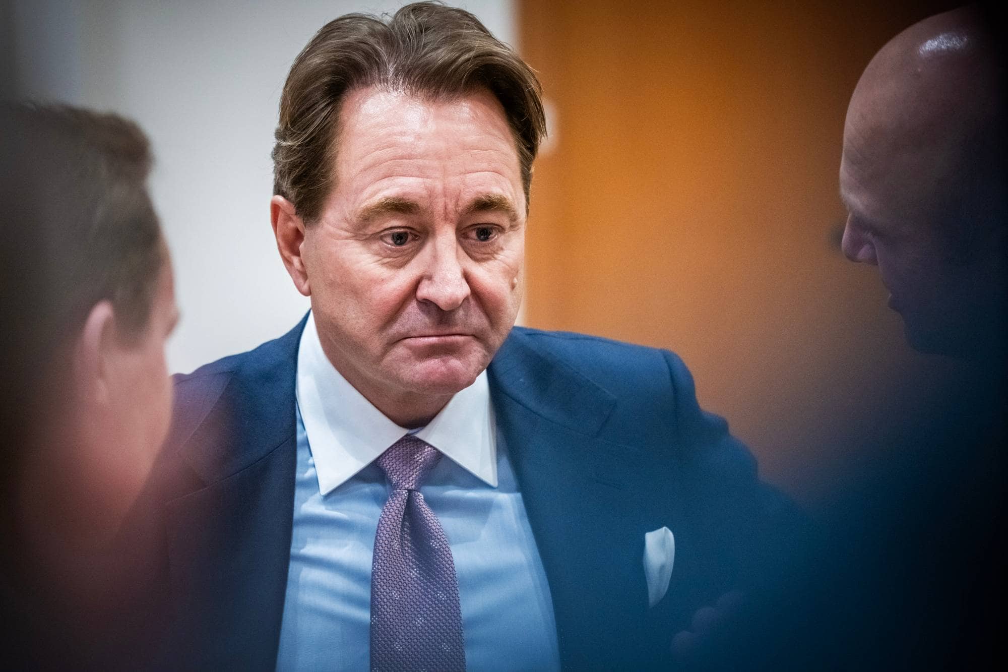 En mann i dress med slips ser ned. To andre menn, hvorav den ene er skallet, er i bakgrunnen. Bakgrunnen er delt i en lys vegg og en oransje vegg. (Bildebeskrivelsen er laget av en KI-tjeneste)