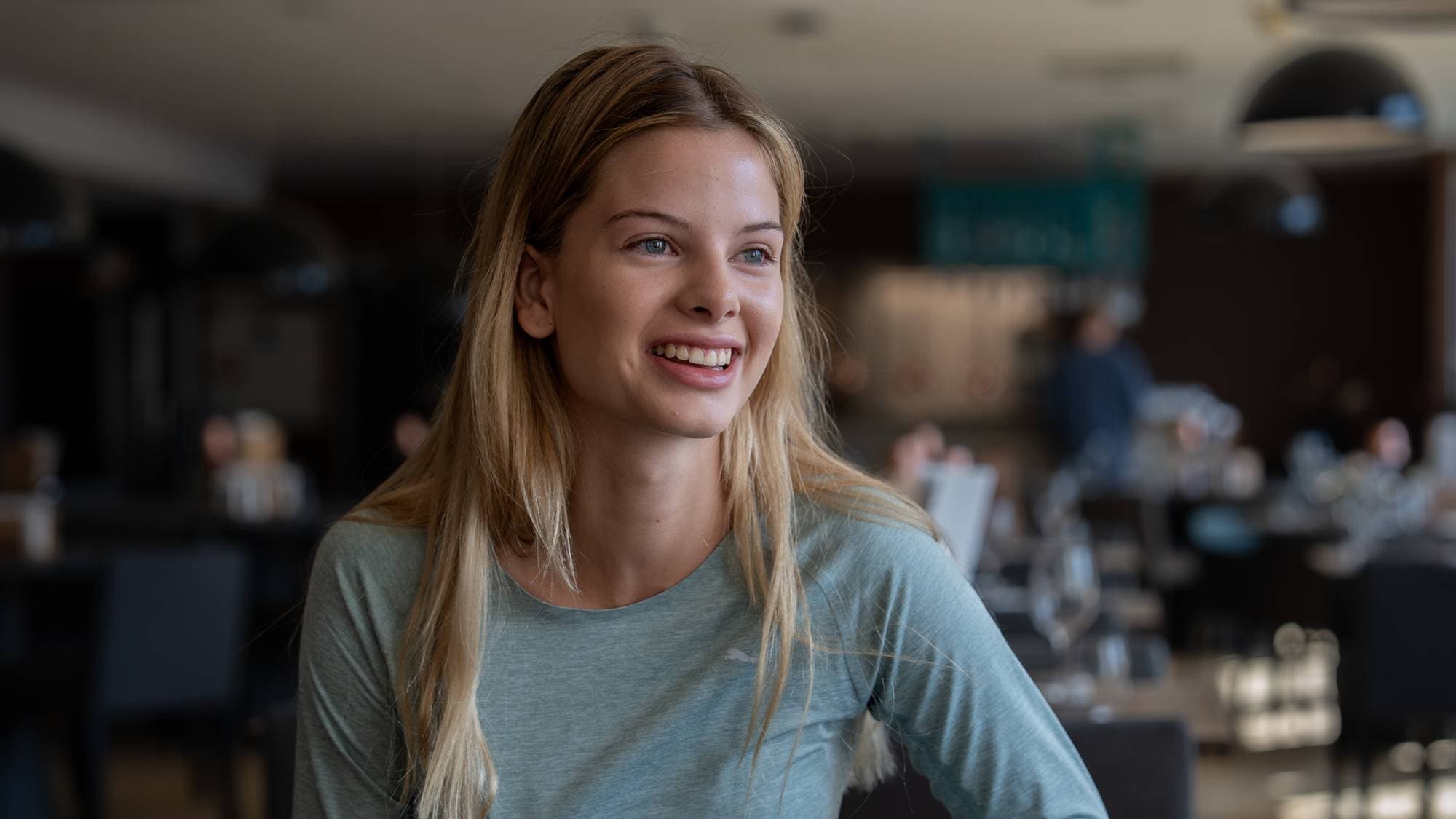 Ung kvinne med langt blondt hår smiler i et restaurantlokale. (Bildebeskrivelsen er laget av en KI-tjeneste)
