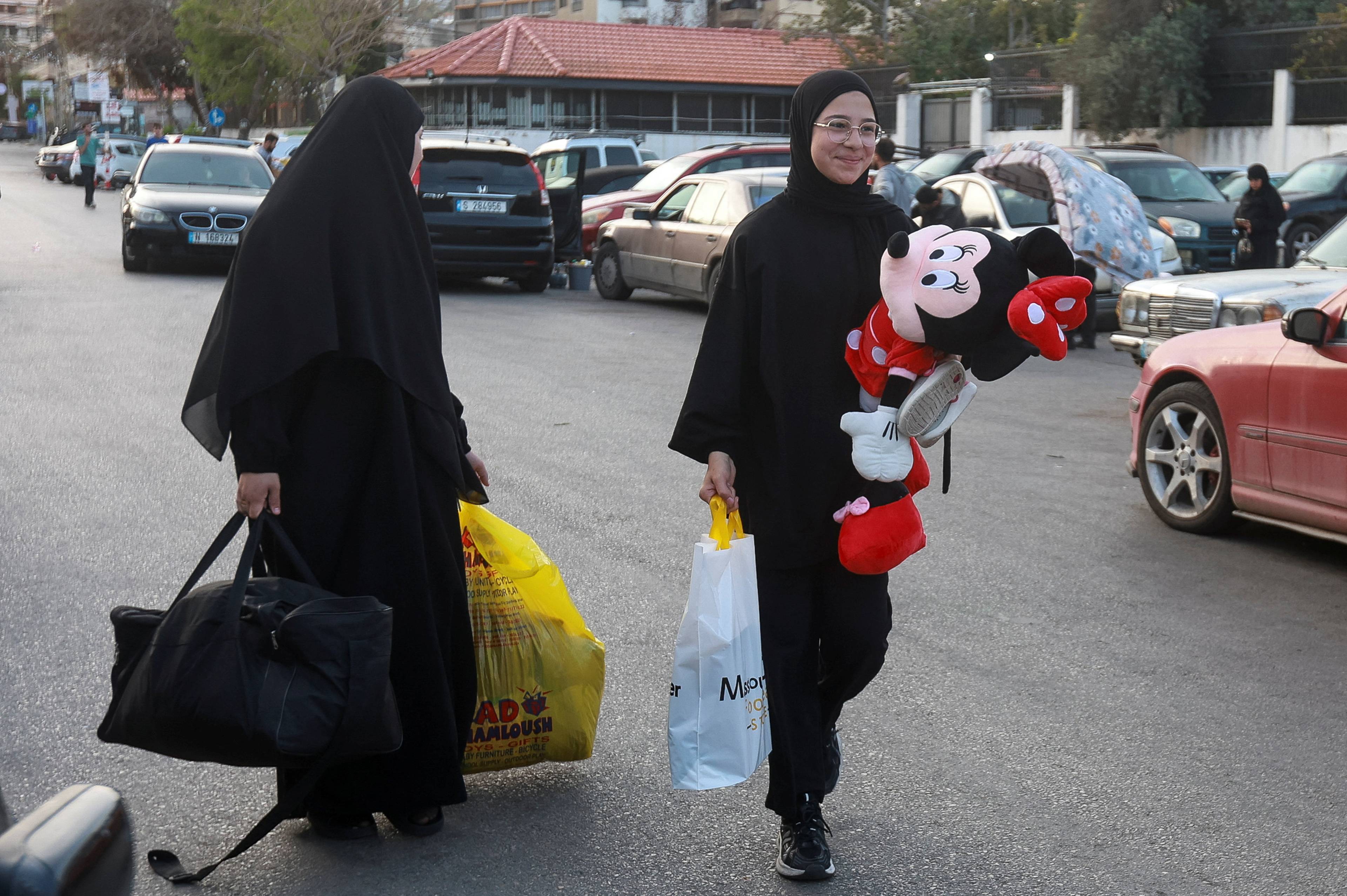 To kvinner i heldekkende klær går på en vei. Den ene bærer en stor svart bag og en gul plastpose. Den andre bærer en hvit pose og en stor bamseformet Mickey Mouse-figur. (Bildebeskrivelsen er laget av en KI-tjeneste)