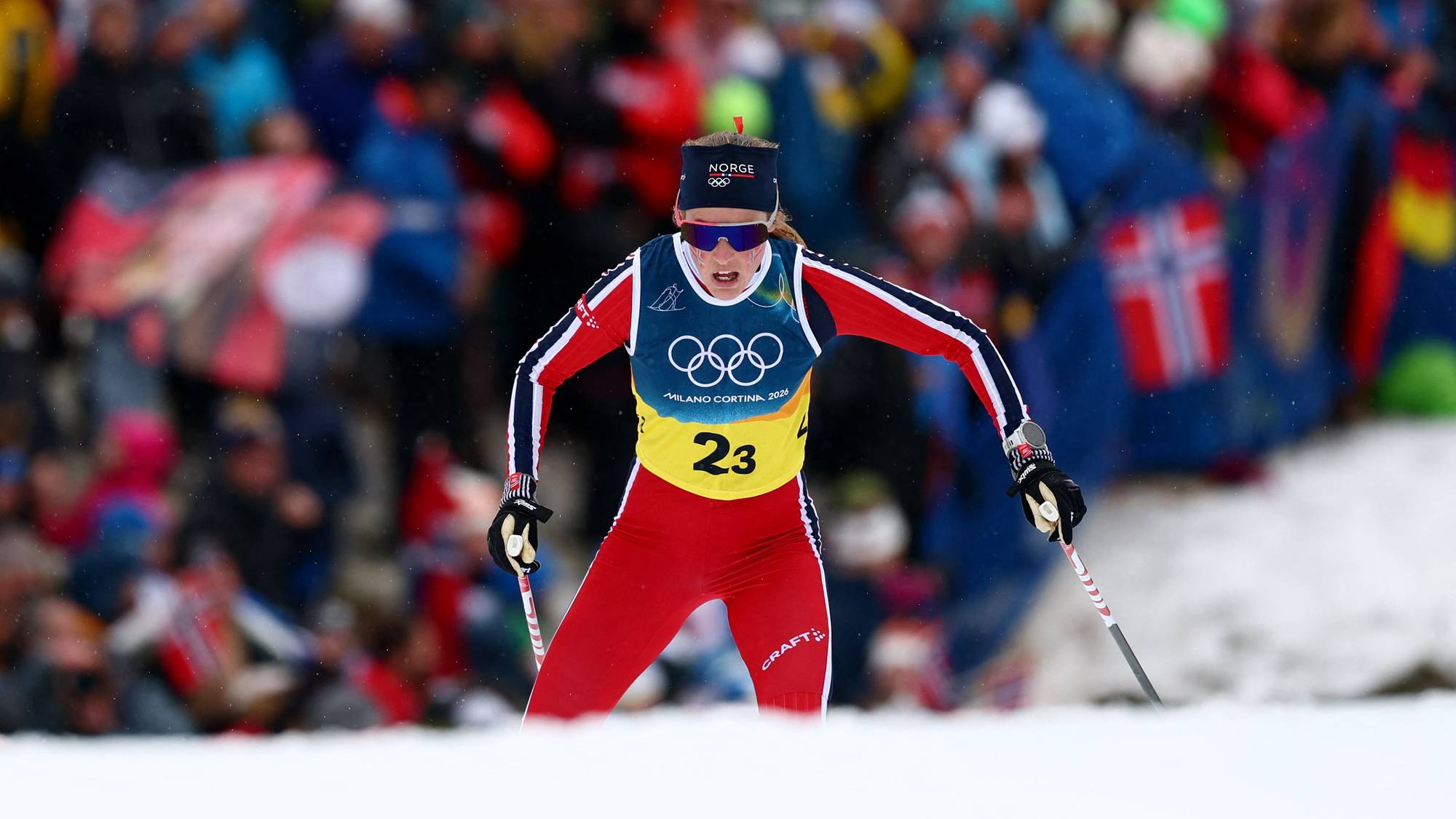 En skiskytter i rød og hvit drakt deltar i et løp. Atleten bærer nummer 23 og har på seg solbriller og pannebånd med teksten "NORGE". Tilskuerne står tett bak et sperrebånd i bakgrunnen. (Bildebeskrivelsen er laget av en KI-tjeneste)