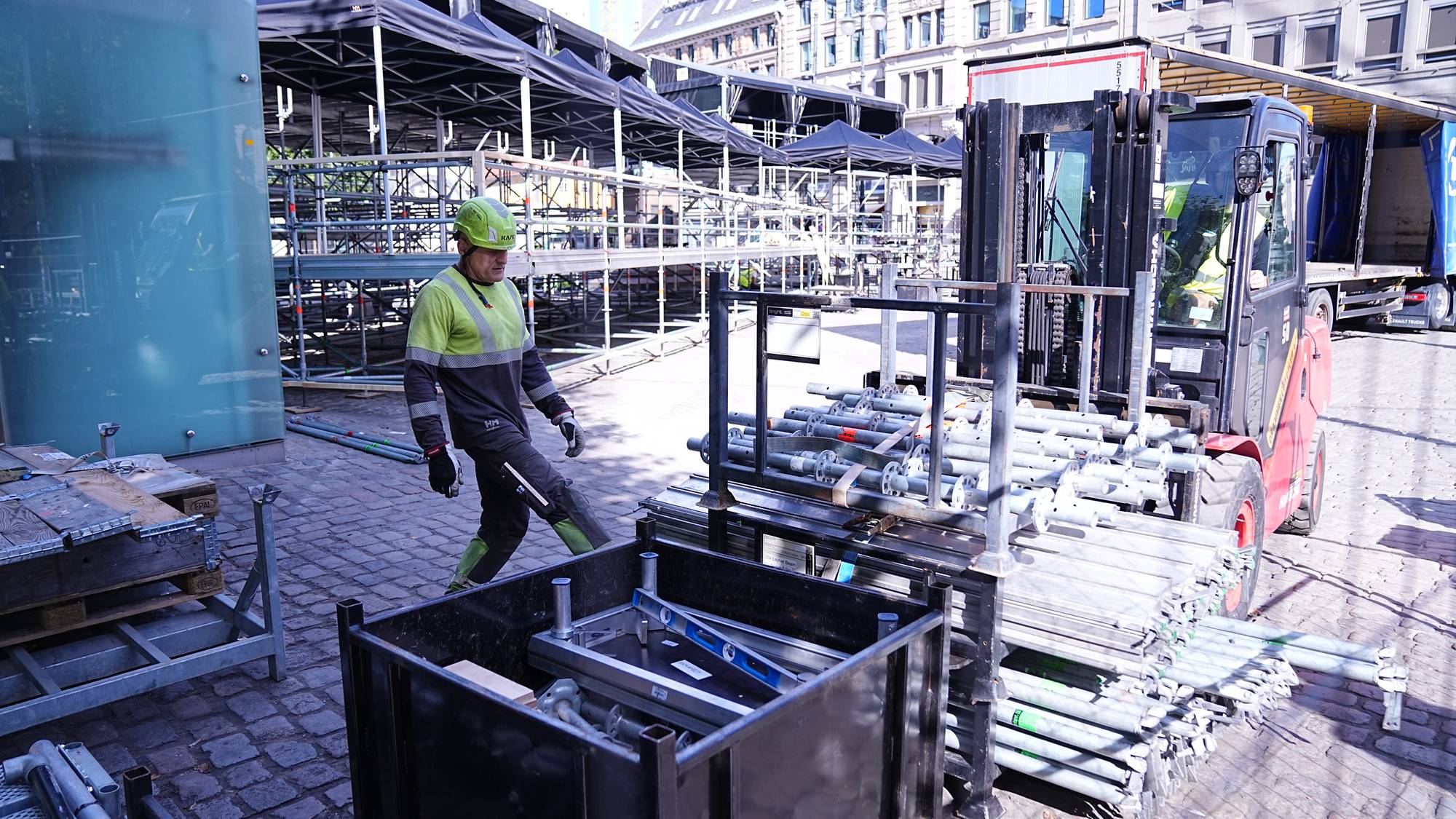 En arbeider i gul refleksvest og hjelm klargjør stillaser ved en gaffeltruck på en byggeplass. (Bildebeskrivelsen er laget av en KI-tjeneste)