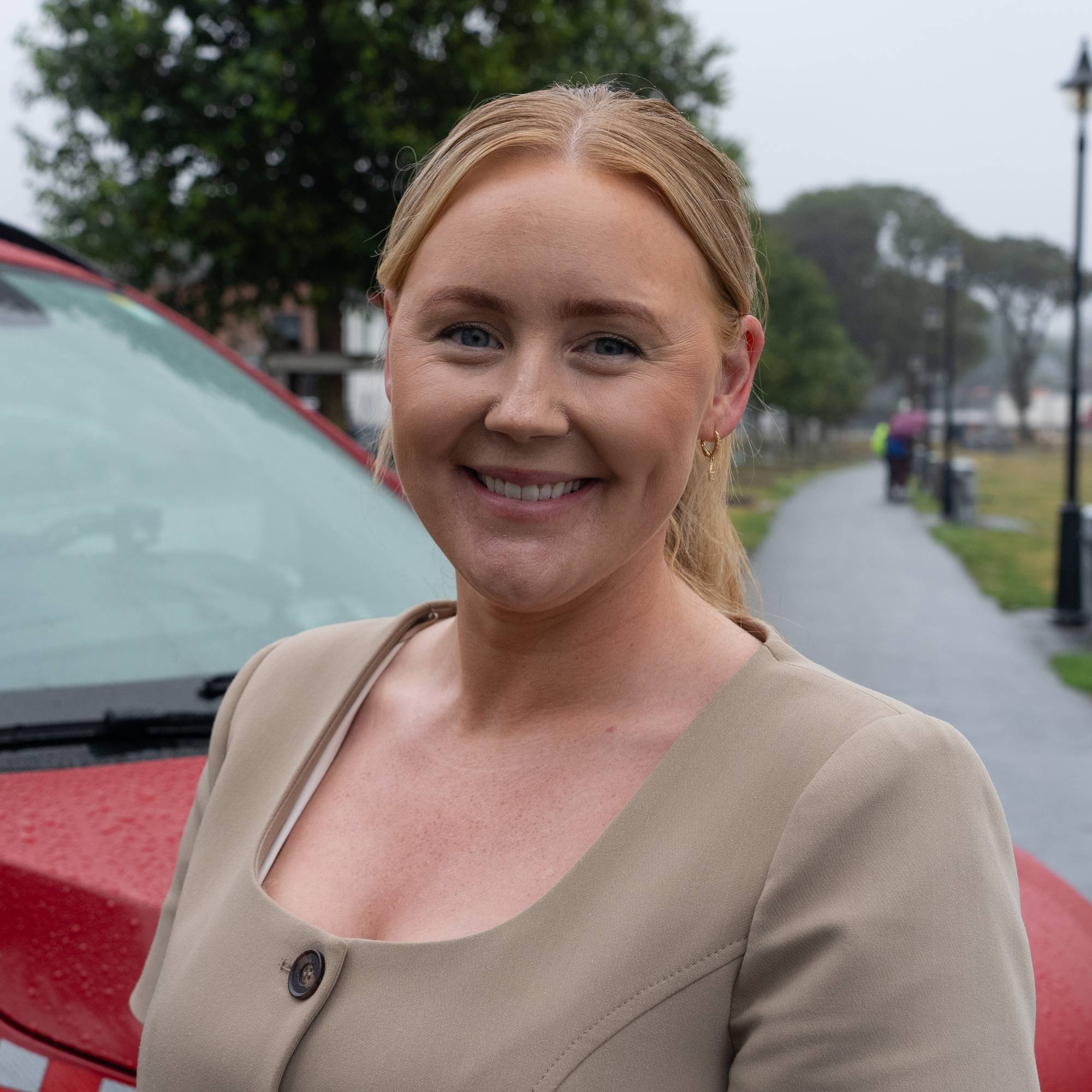 En smilende kvinne foran en rød utrykningsbil utendørs. (Bildebeskrivelsen er laget av en KI-tjeneste)