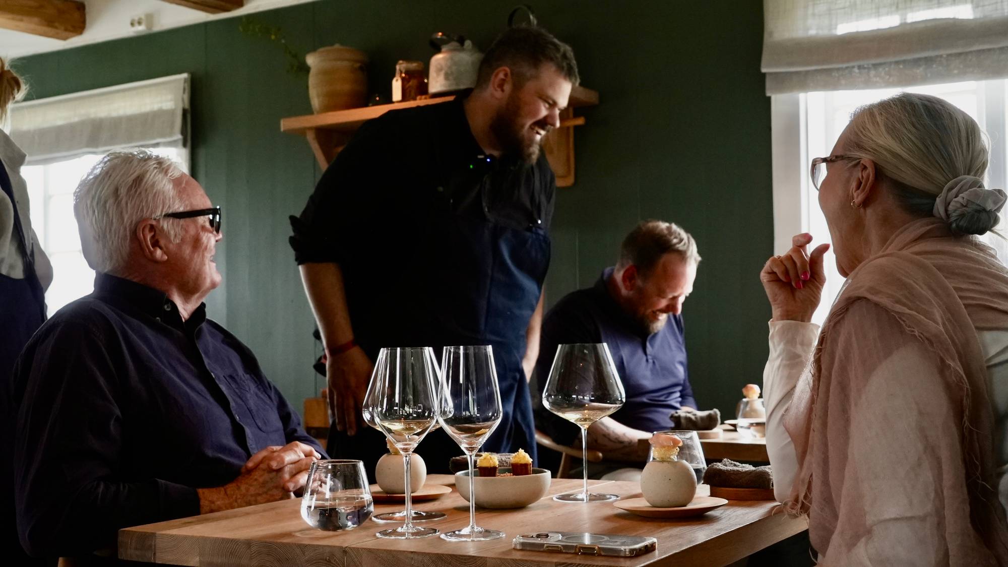 En kokk serverer mat til gjester ved et langbord. De sitter ved et dukket bord med glass og småretter. Rommet har en mørkegrønn vegg og synlige takbjelker. (Bildebeskrivelsen er laget av en KI-tjeneste)