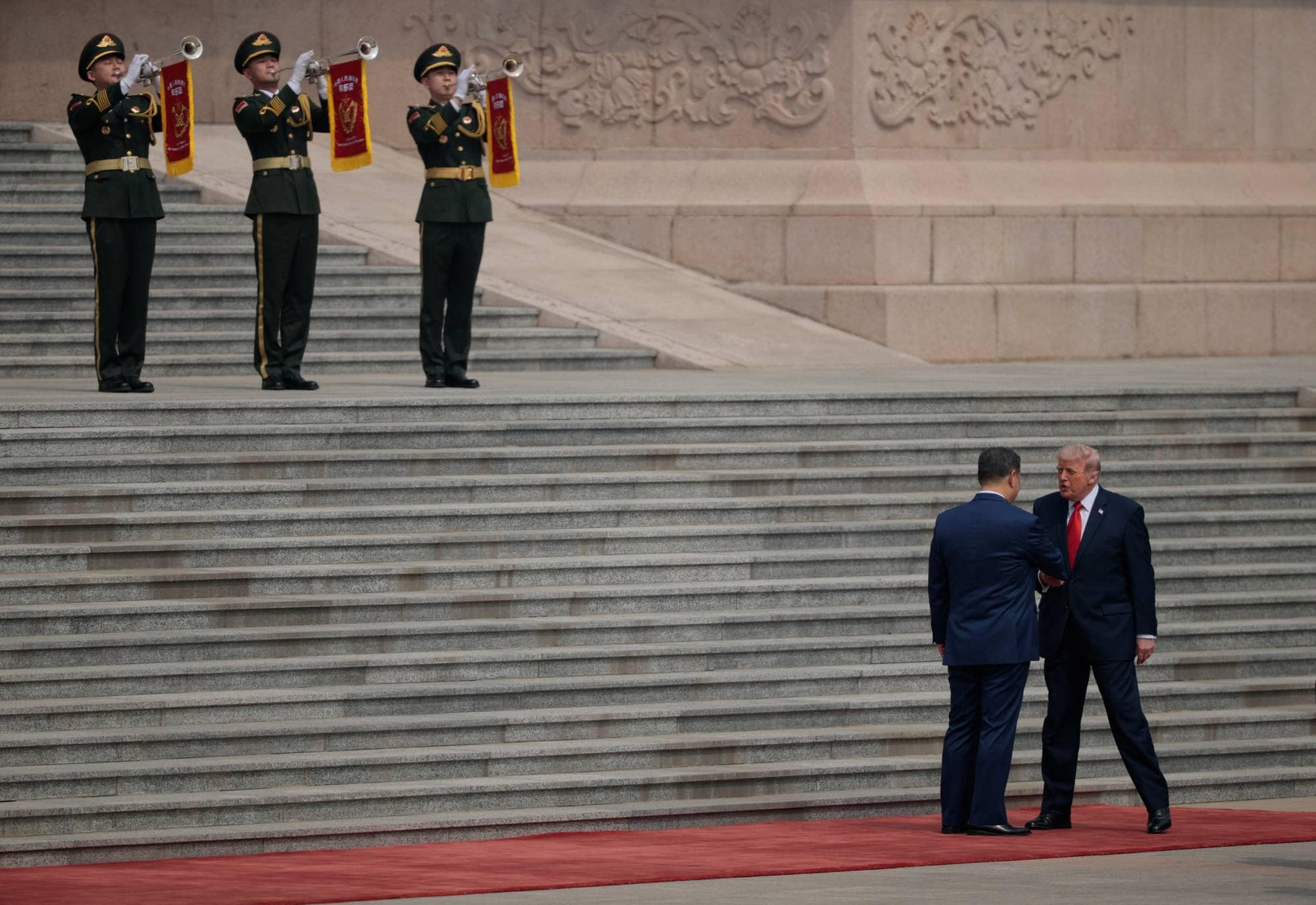 To kinesiske soldater spiller trompet. USAs president Donald Trump og Kinas president Xi Jinping håndhilser. De står på et rødt teppe foran en stor steintrapp.