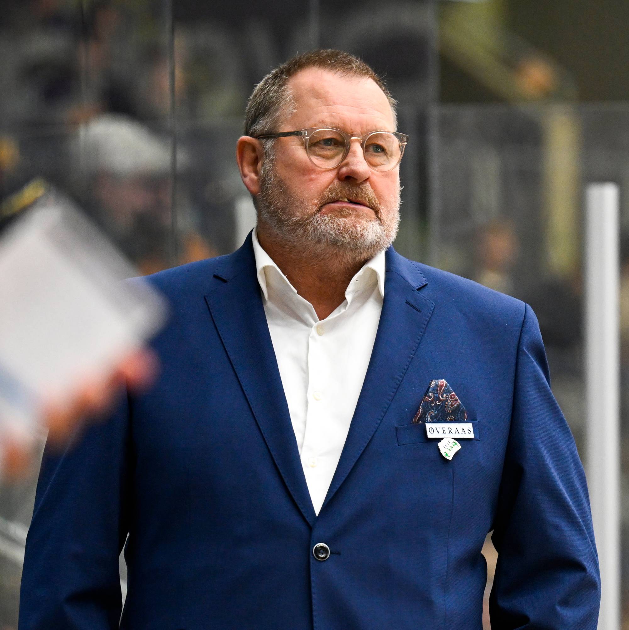 Ishockeytrener i blå dress står ved vantet i en ishall med spillere i forgrunnen. 