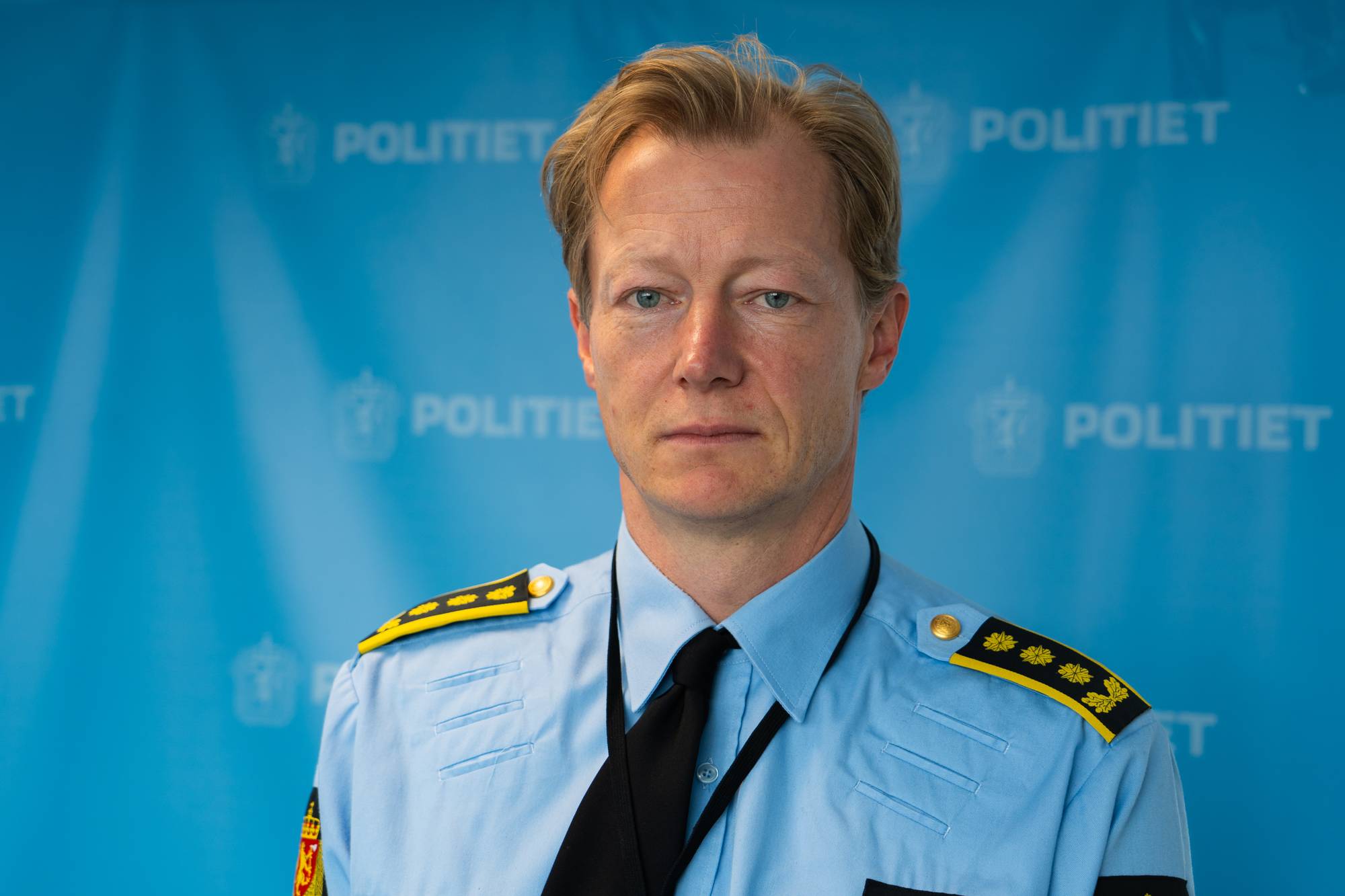 Portrett av politimann med blondt hår og uniform framfor ein blå pressevegg. 