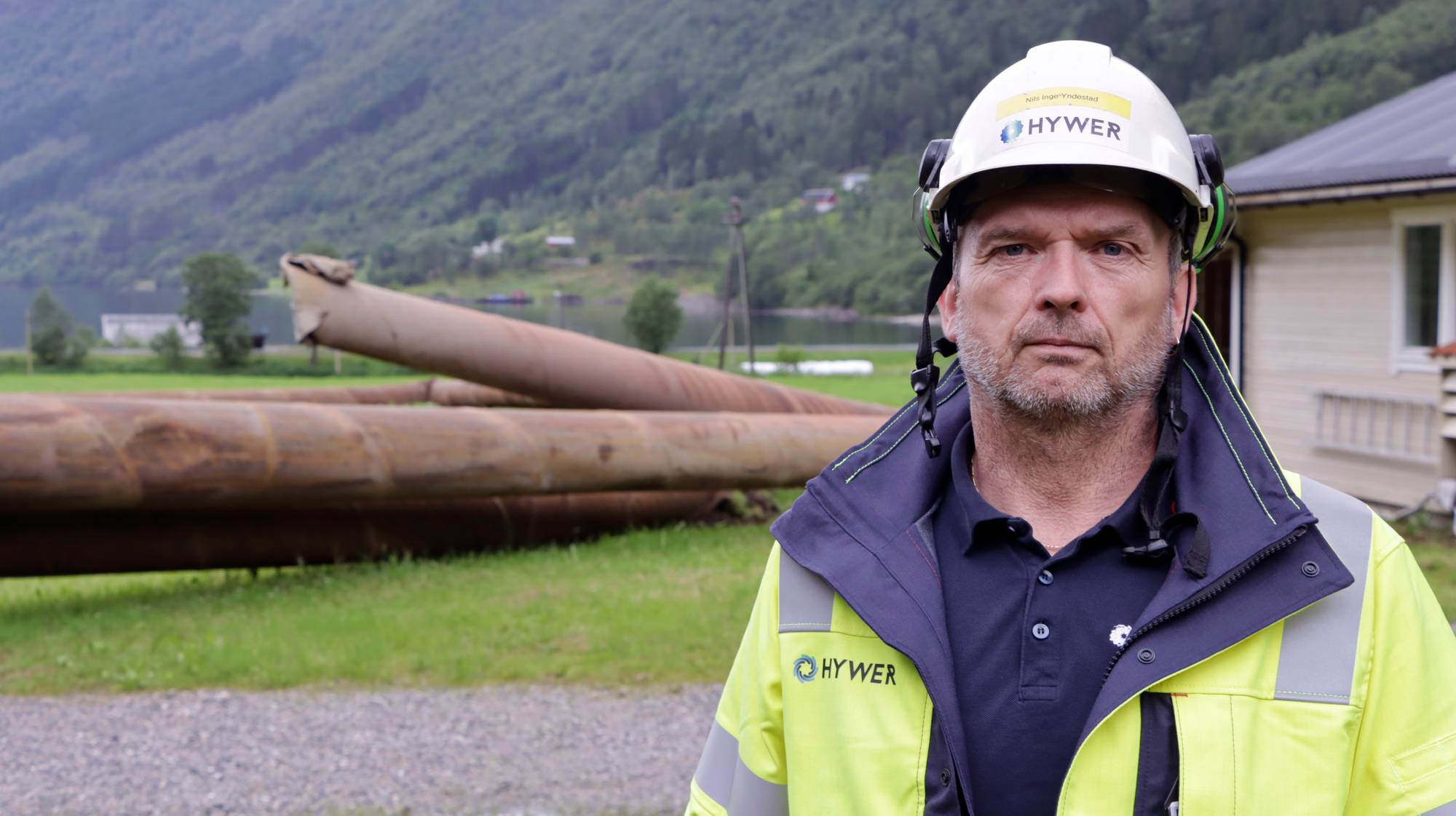 Mann med vernehjelm foran store rør på et anleggsområde ved en fjord. (Bildebeskrivelsen er laget av en KI-tjeneste)