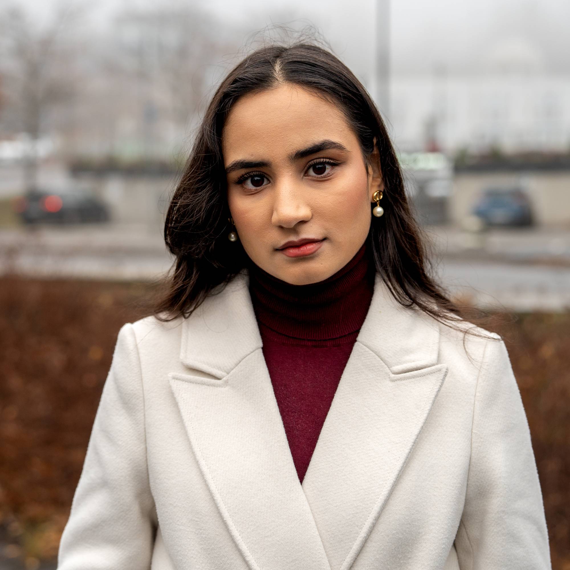En person står foran en utydelig bakgrunn på en tåkete dag. Personen har på seg en lys, strukturert frakk og en mørk genser. Omgivelsene inkluderer trær med visne blader og biler som kjører i bakgrunnen. Det er tydelig at det er tidlig vår, med en dempet fargepalett i landskapet.