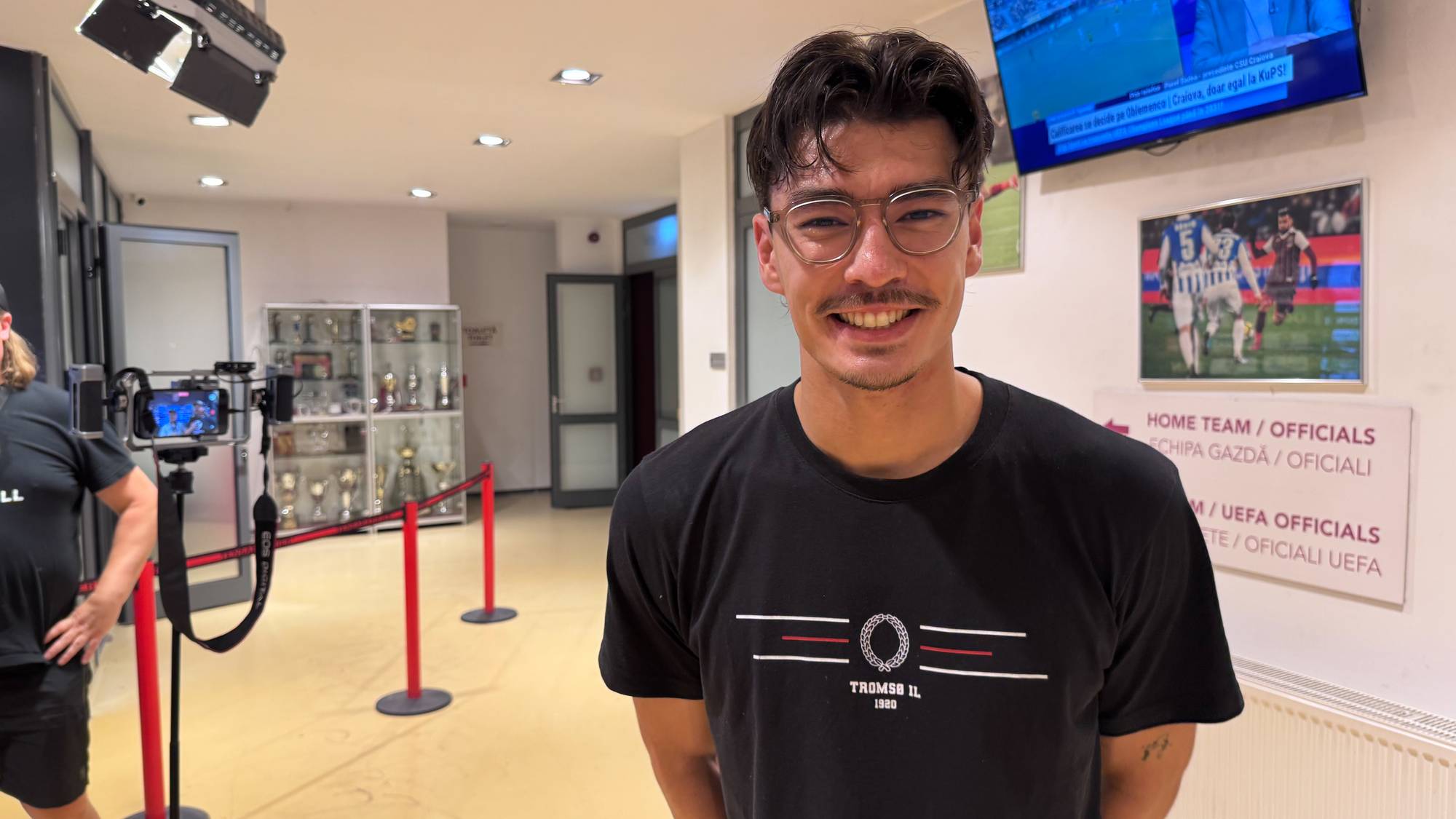 Smilende mann i Tromsø IL-t-skjorte i et korridorområde med TV-skjermer og kamerautstyr. (Bildebeskrivelsen er laget av en KI-tjeneste)