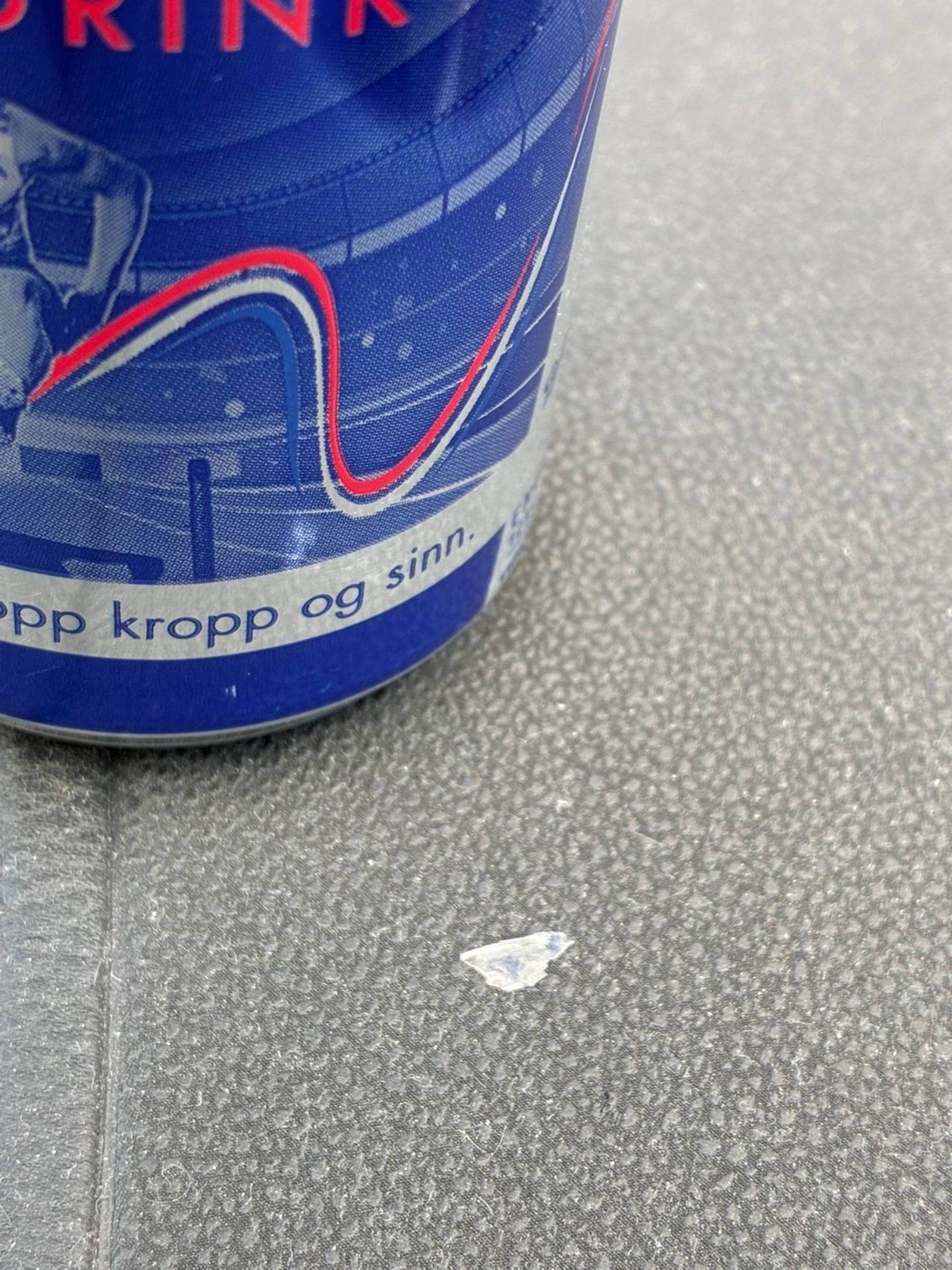 Nærbilde av en blå energidrikkboks stående på et grått underlag med en liten glassbit ved siden. (Bildebeskrivelsen er laget av en KI-tjeneste)