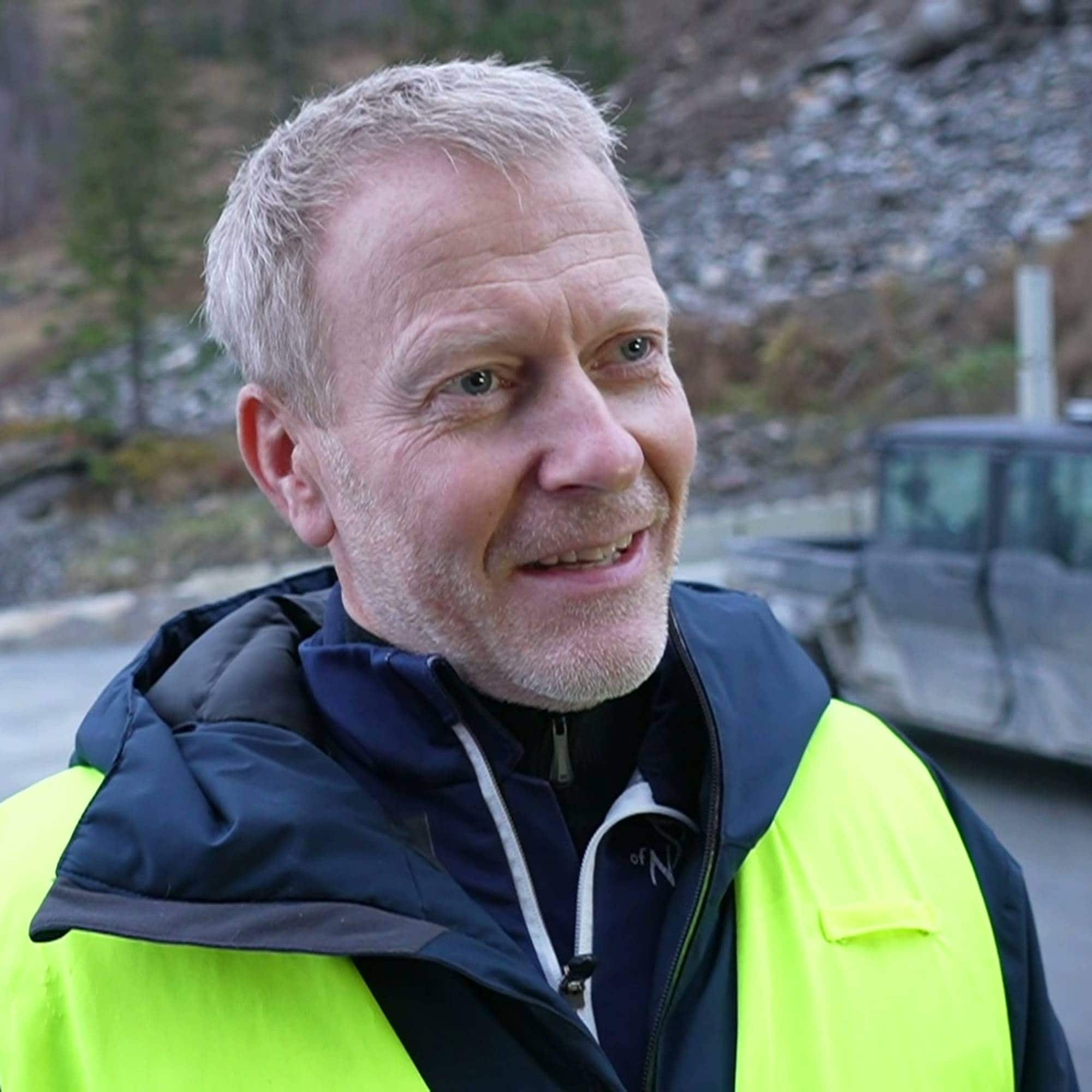 Mann med grått hår i gul refleksvest står utendørs ved en parkeringsplass. 
