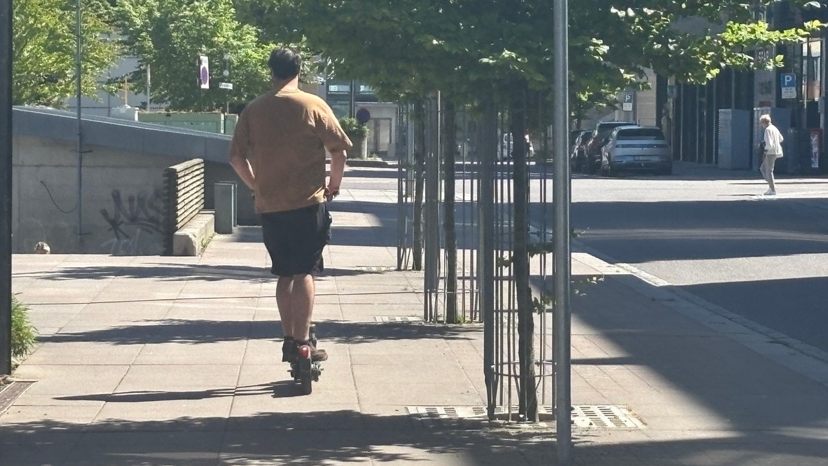 Person i beige t-skjorte kjører elektrisk sparkesykkel på fortauet i en bygate. FOTO: Maya Bråthen/NRK