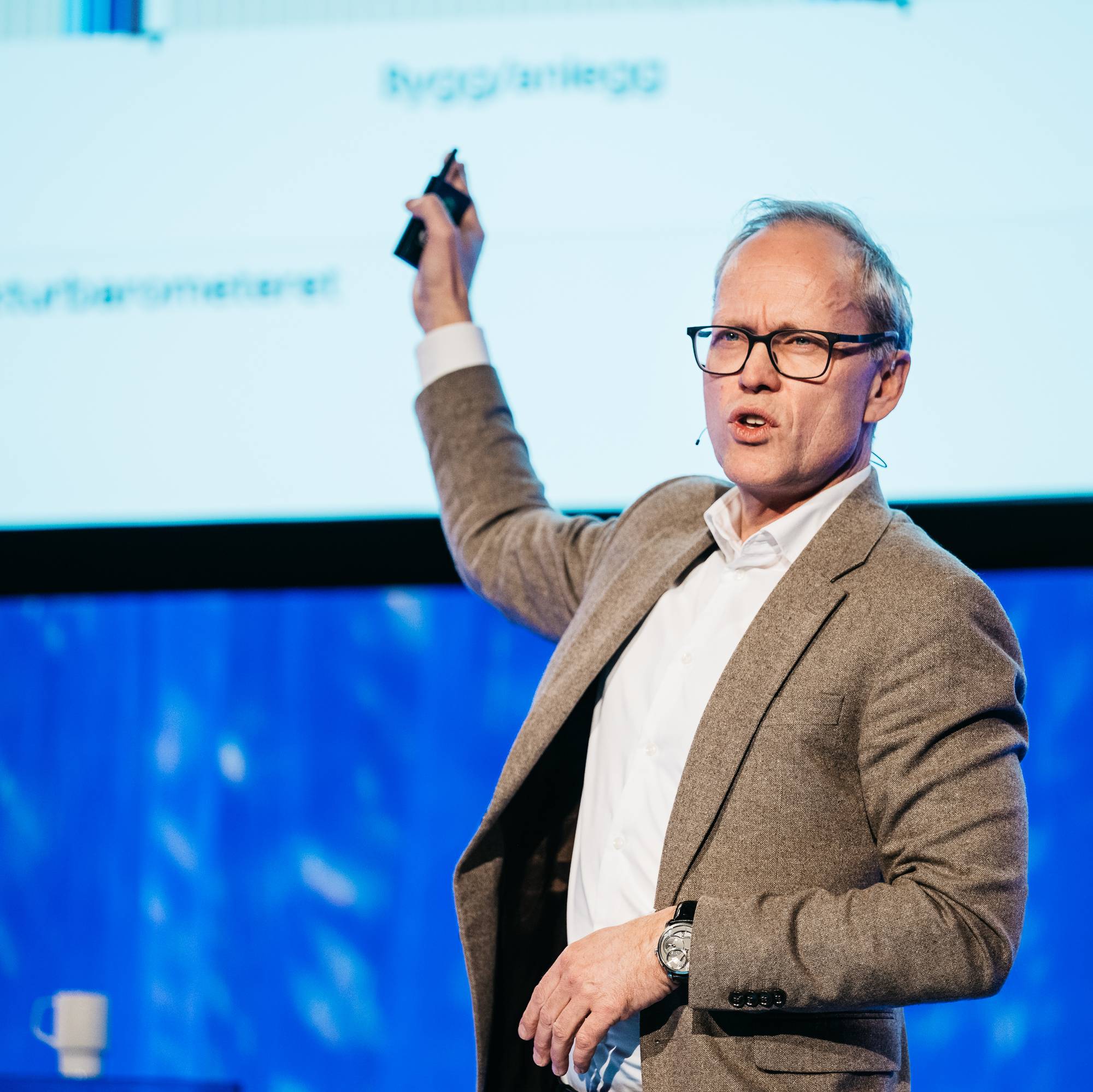 En mann holder en klikker og peker opp mot en skjerm med grafer. Han har på seg en blazer og briller. Bak ham er en stor skjerm med tekst og grafer. Mannen snakker til et publikum. (Bildebeskrivelsen er laget av en KI-tjeneste)