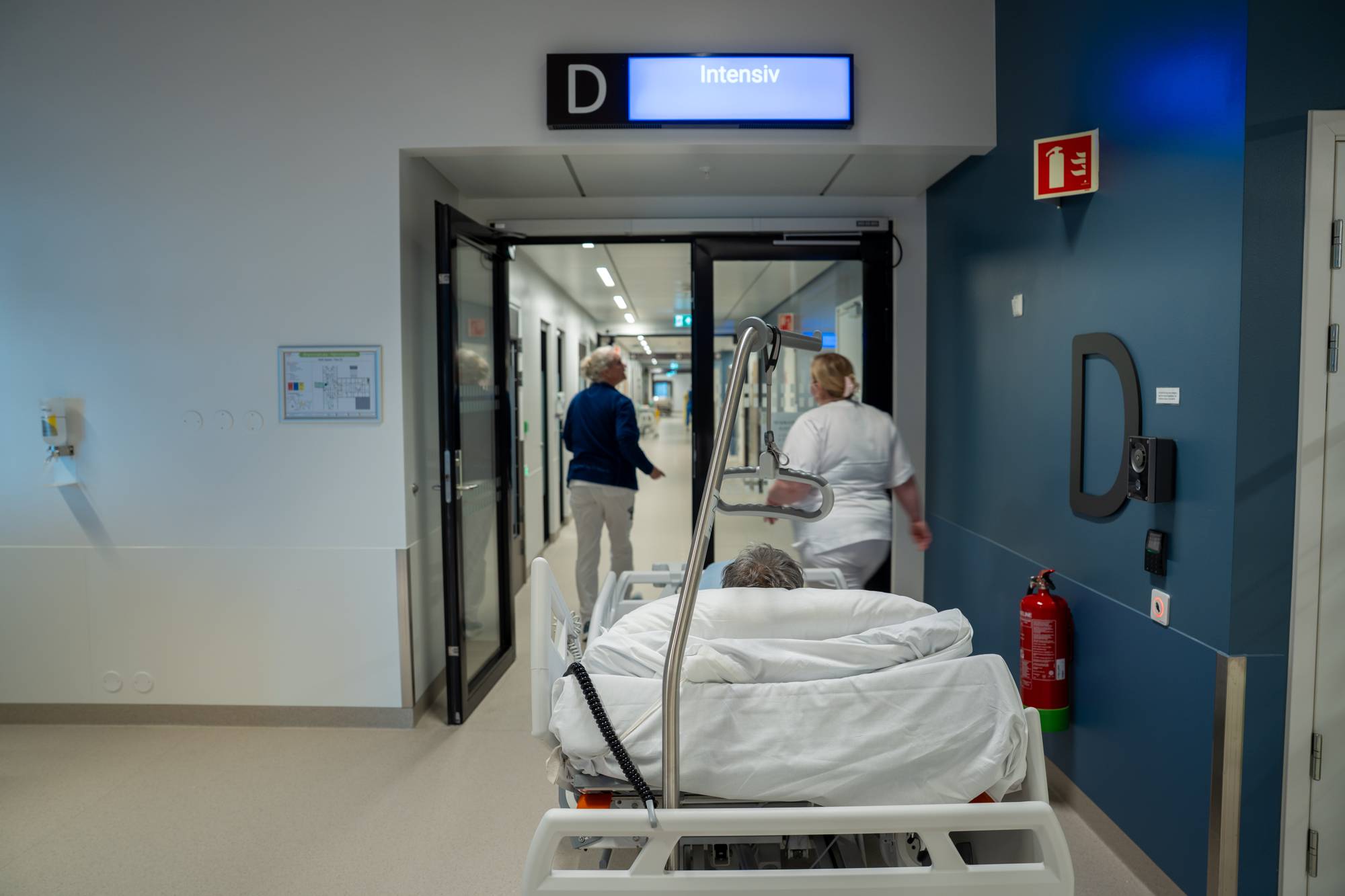 Bildet viser en person som ligger i en sykehusseng, med fokus på intensivavdelingen. I bakgrunnen går en sykepleier og en besøkende mot inngangen til avdelingen. Omgivelsene er moderne og lyssterke, med klare skilt og en ren, lukket korridor. Det er synlig en brannslukker på veggen nær inngangen. (Bildebeskrivelsen er laget av en KI-tjeneste)