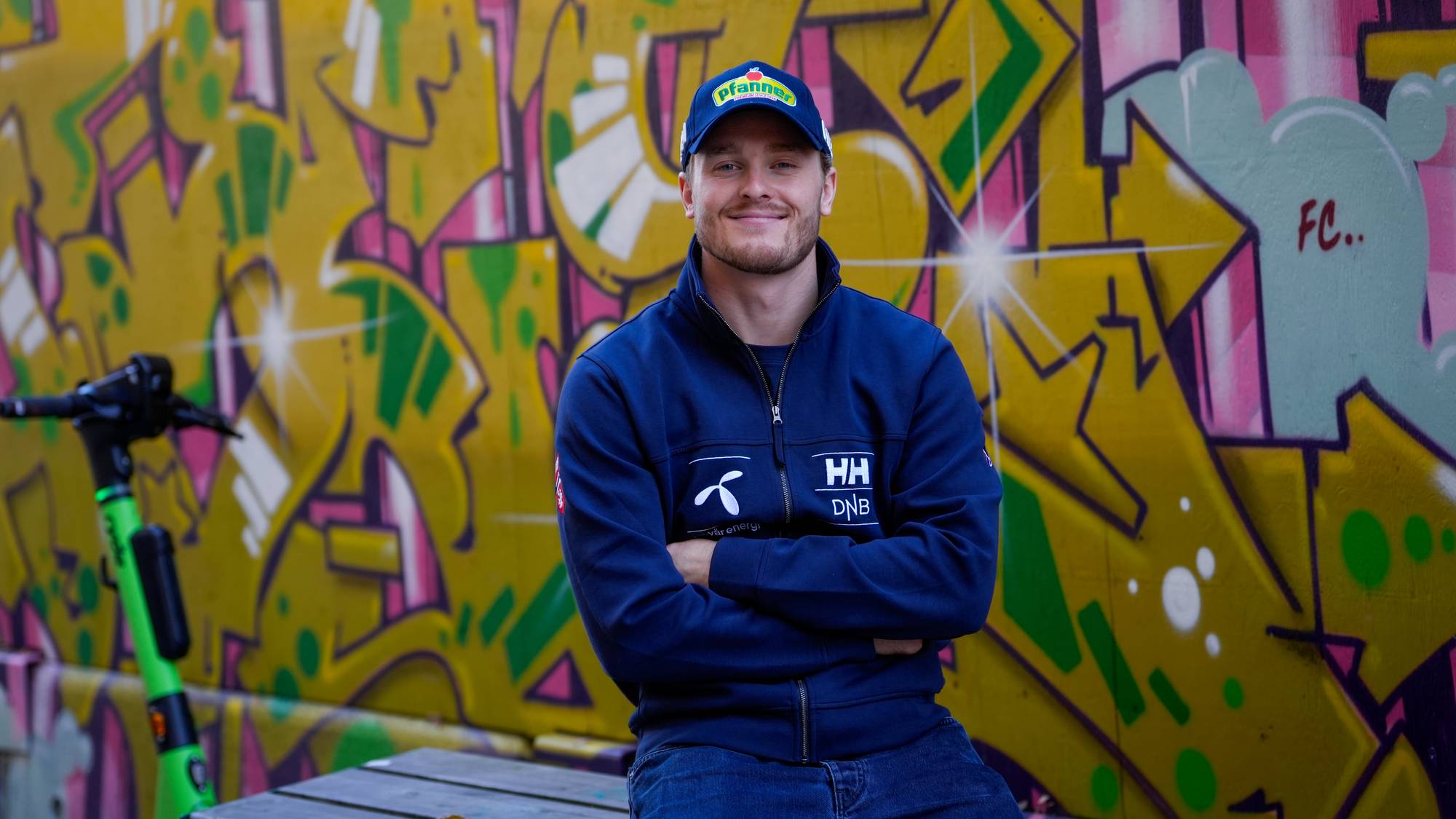 En mann sitter foran en graffitivegg med en elsparkesykkel ved siden av seg. (Bildebeskrivelsen er laget av en KI-tjeneste)