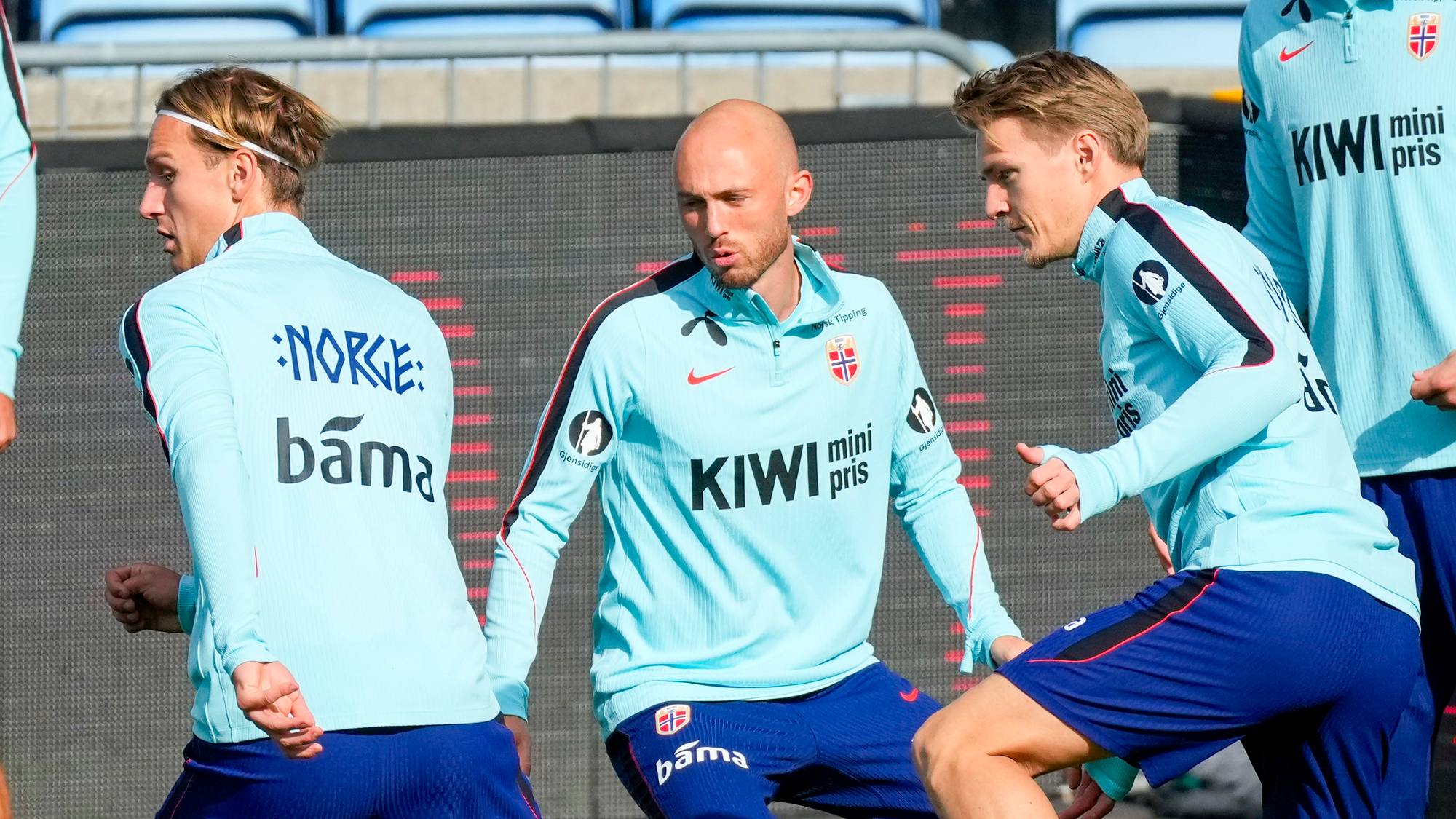 Norske landslagsspillere i fotball trener på gressbane foran tomme tribuner. (Bildebeskrivelsen er laget av en KI-tjeneste)