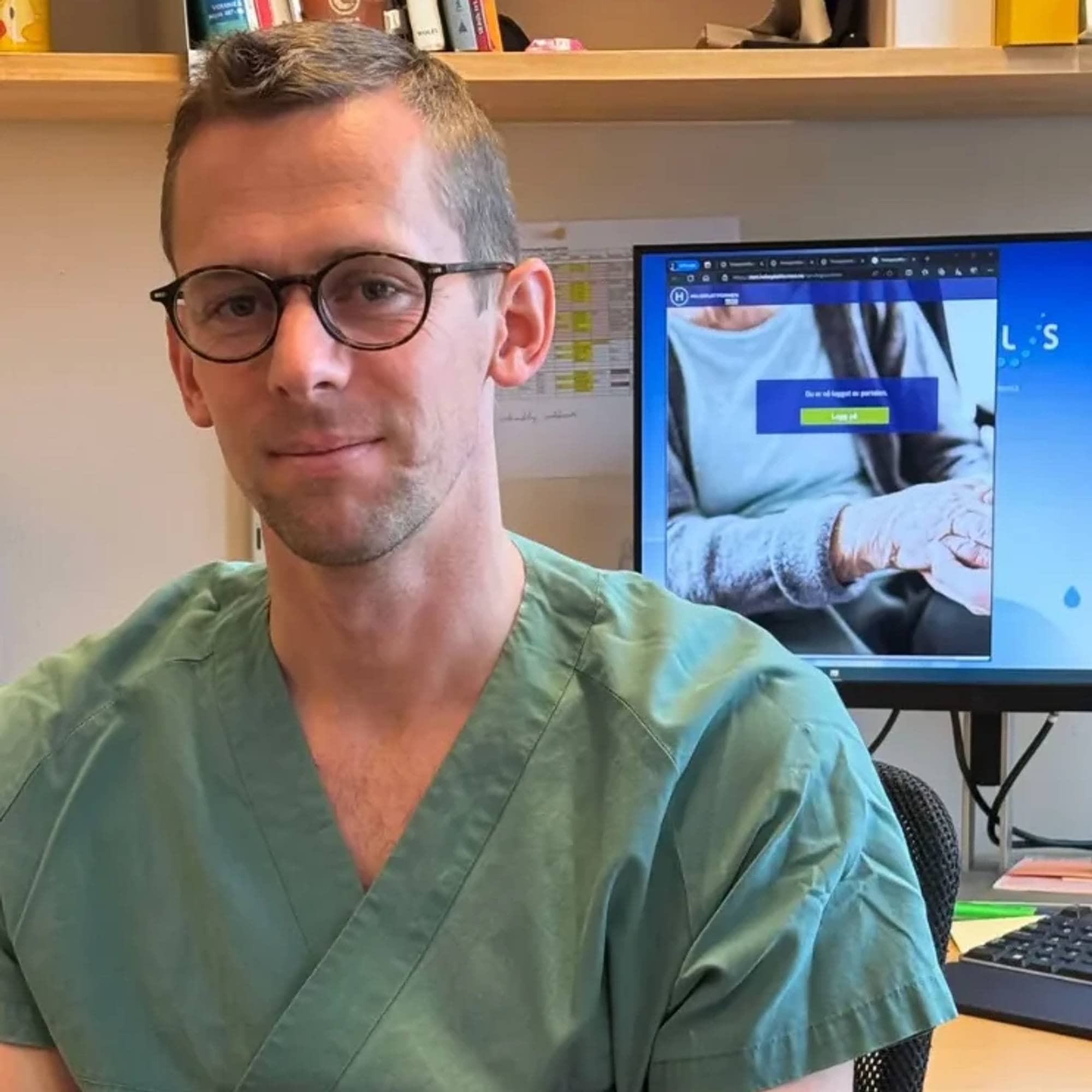 Forsker og lege sitter med grønn uniform på en kontrostol. Han har briller og kort hår.