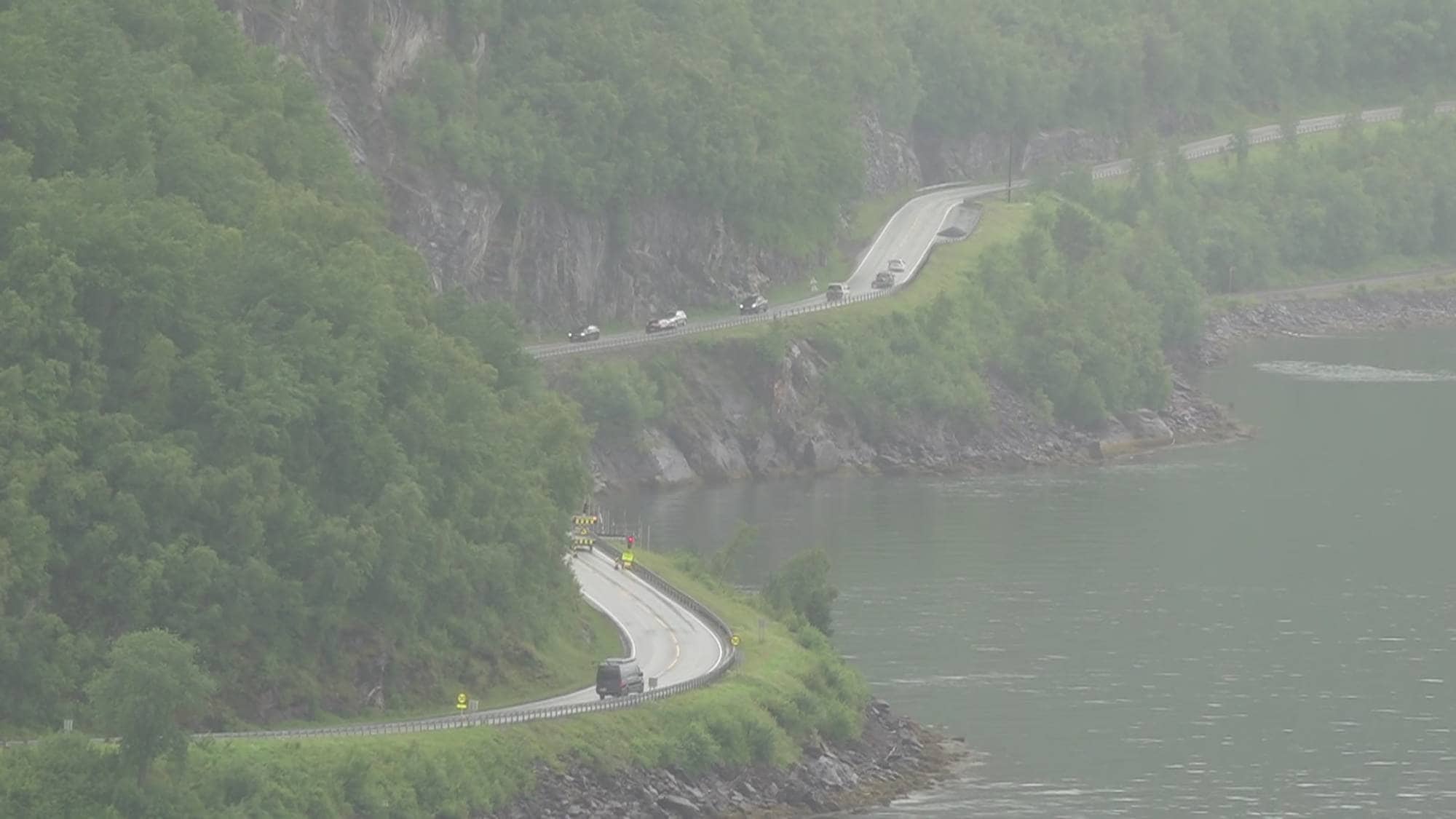 Biler kjører på en svingete vei langs en fjord omgitt av bratte, skogkledde fjellsider i disig vær. (Bildebeskrivelsen er laget av en KI-tjeneste)