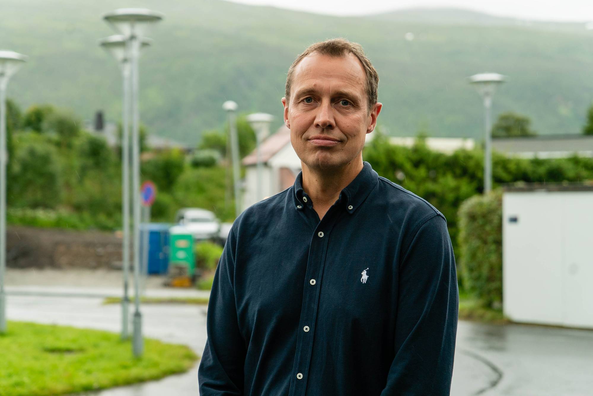 Mann i mørk skjorte står utendørs med grønne omgivelser og fjell i bakgrunnen. (Bildebeskrivelsen er laget av en KI-tjeneste)