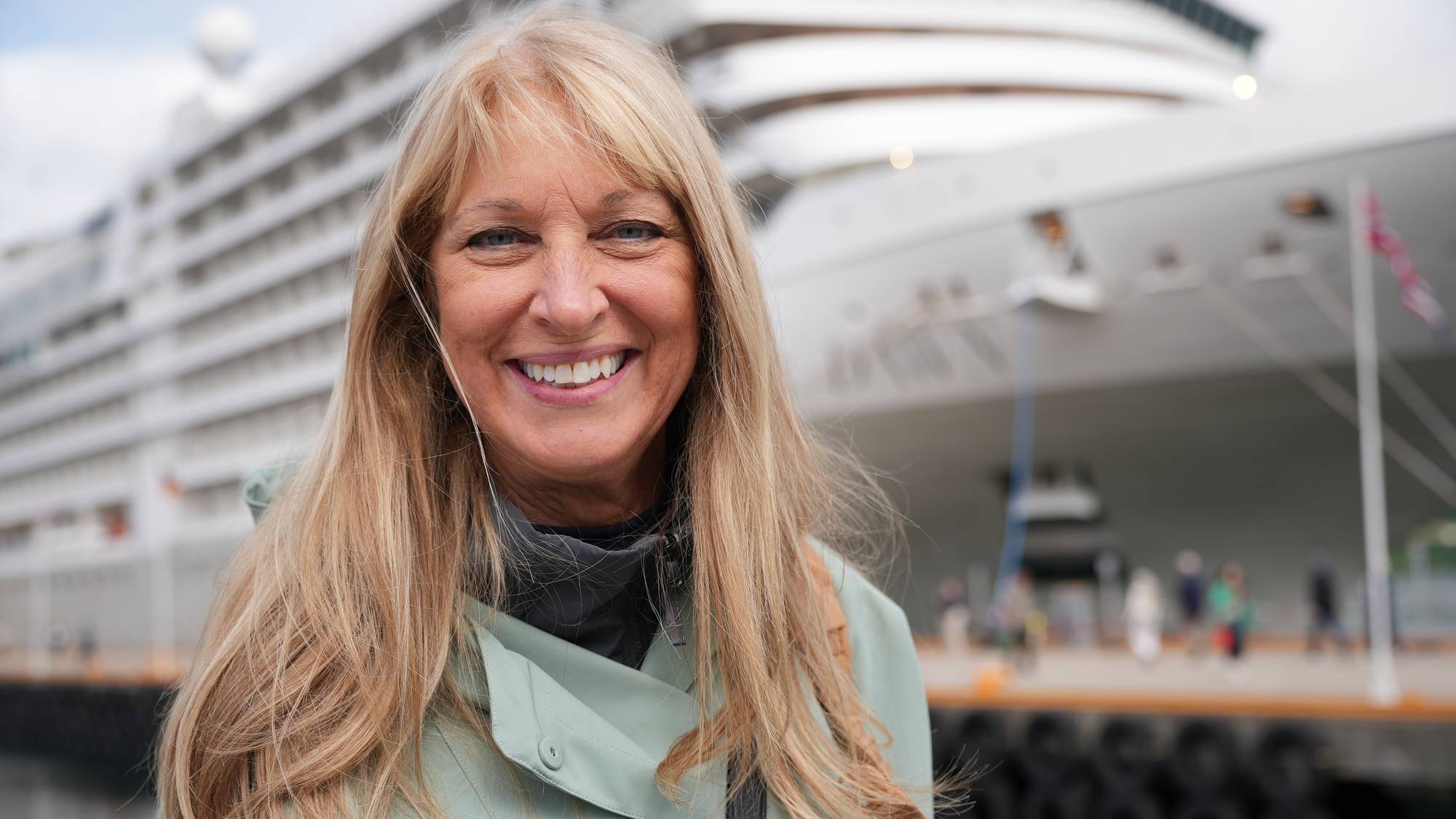 En kvinne med blondt hår smiler mot kameraet. Bak henne ligger et stort cruiseskip fortøyd ved kai. Bakgrunnen er uskarp. (Bildebeskrivelsen er laget av en KI-tjeneste)