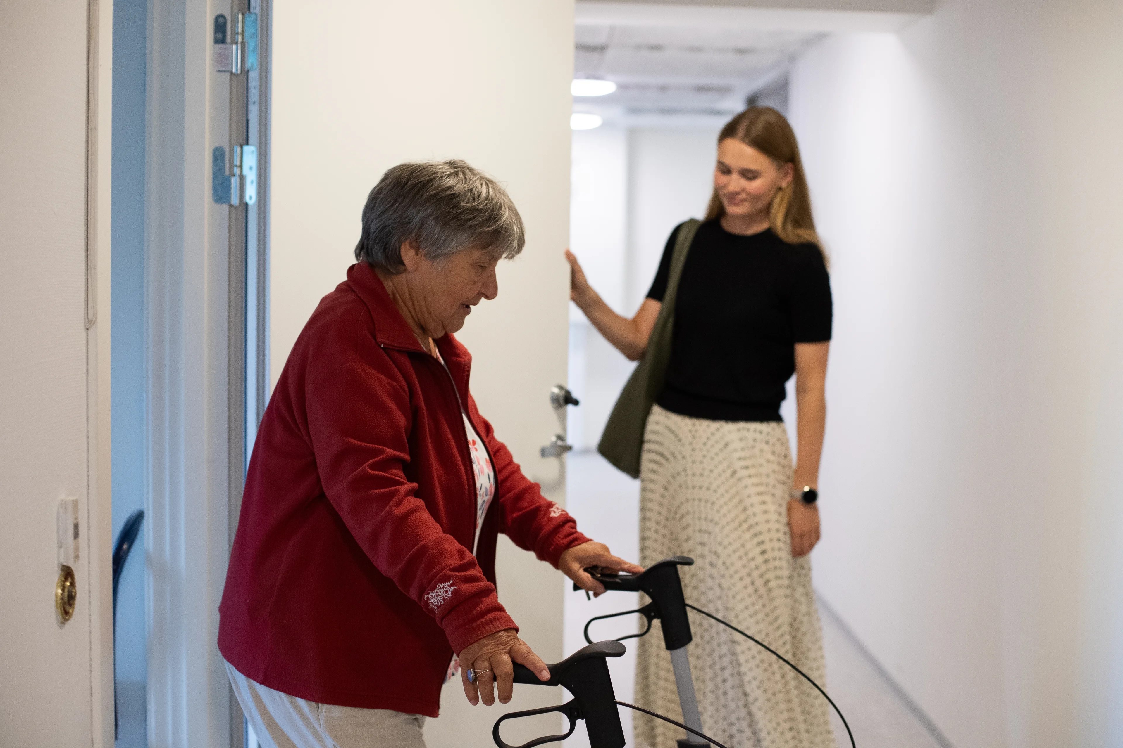 en eldre kvinne med rullator går ut en dør, som en yngre kvinne holder åpen
