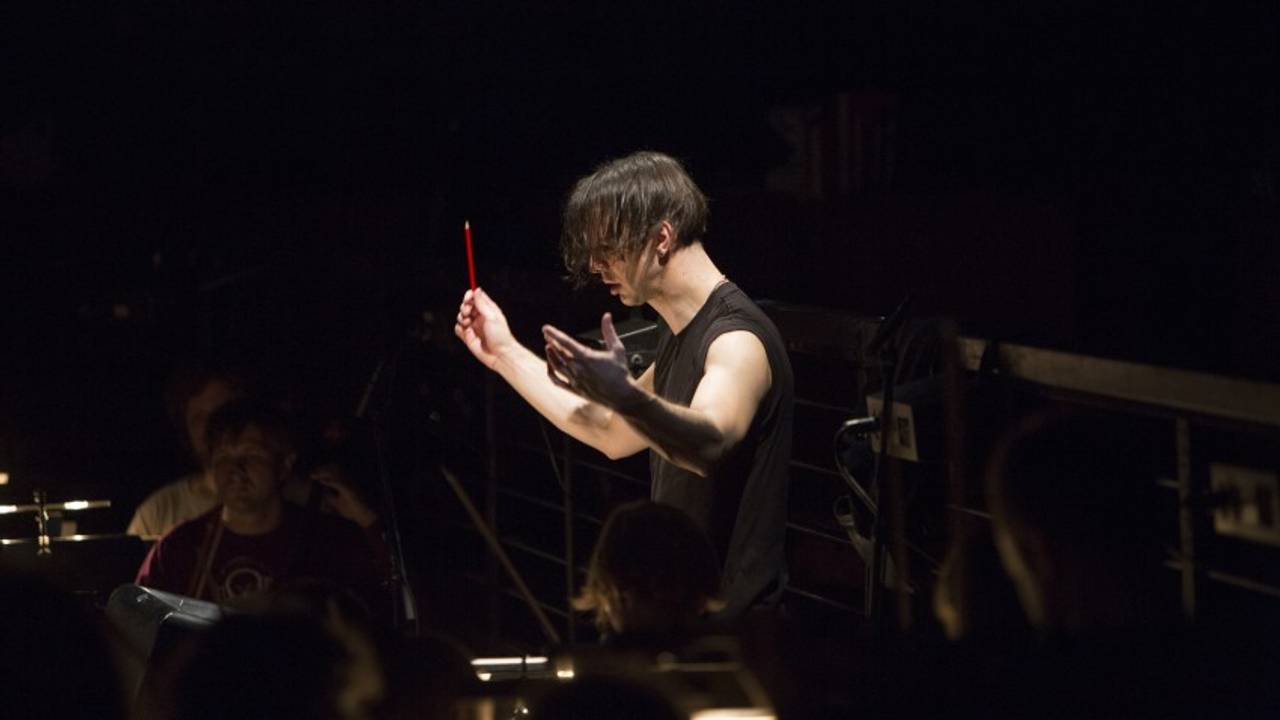 Teodor Currentzis dirigerer. Siett fra siden, ansiktet forover, håret henger forover. Iført ermeløs svart t-skjorte. Musikere i orkesteret skimtes rundt ham i mørket.